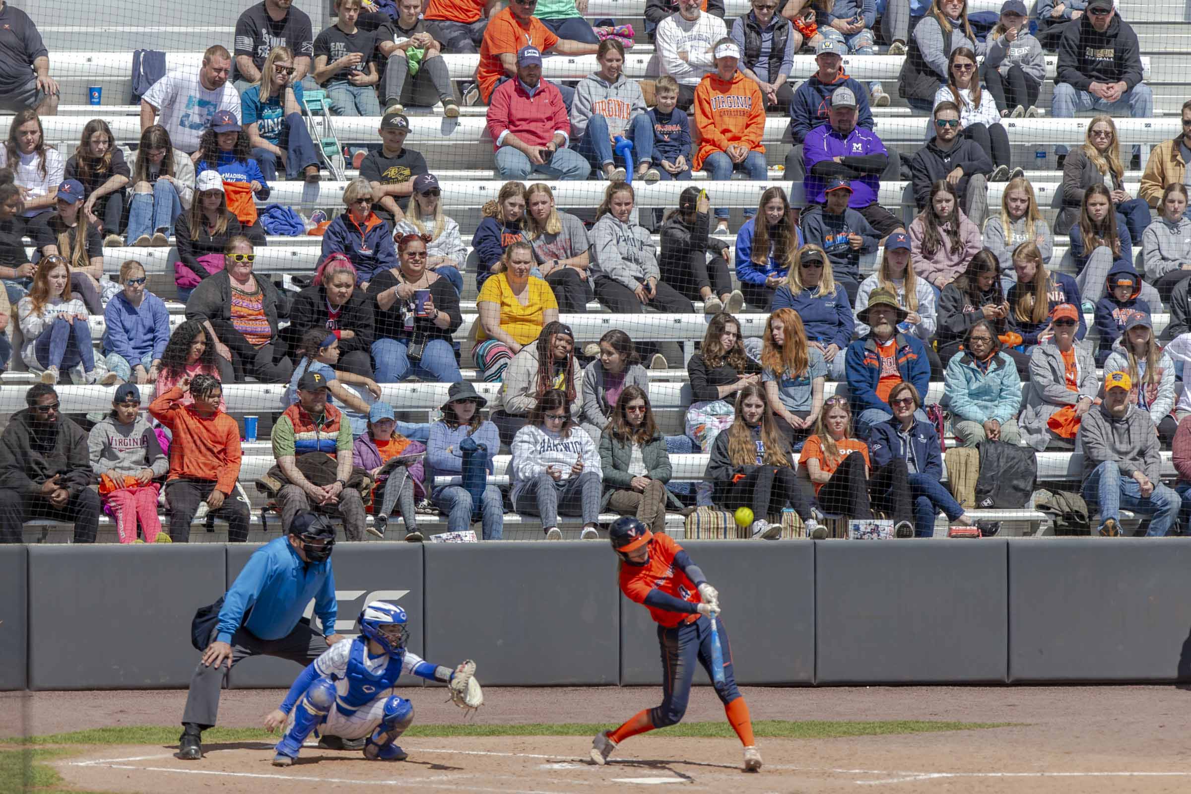 A UVA softball player at bat, hits the ball. Fans look on from the crowded bleachers.