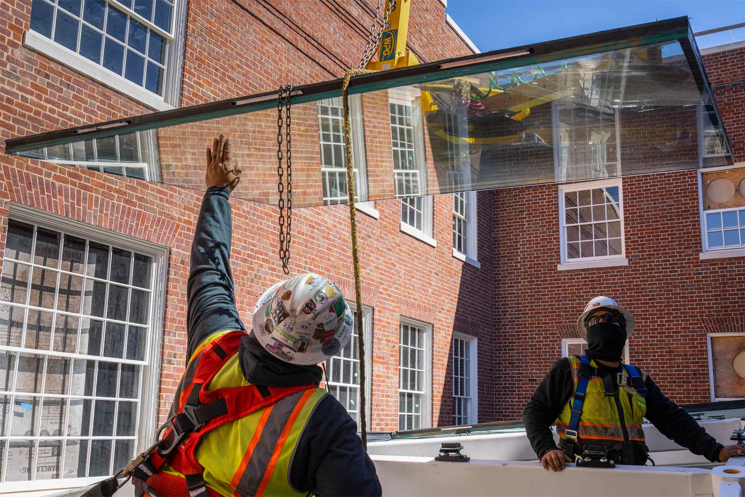 Standing inside the frame, two workers oversee another pane of glass as the crane lowers it