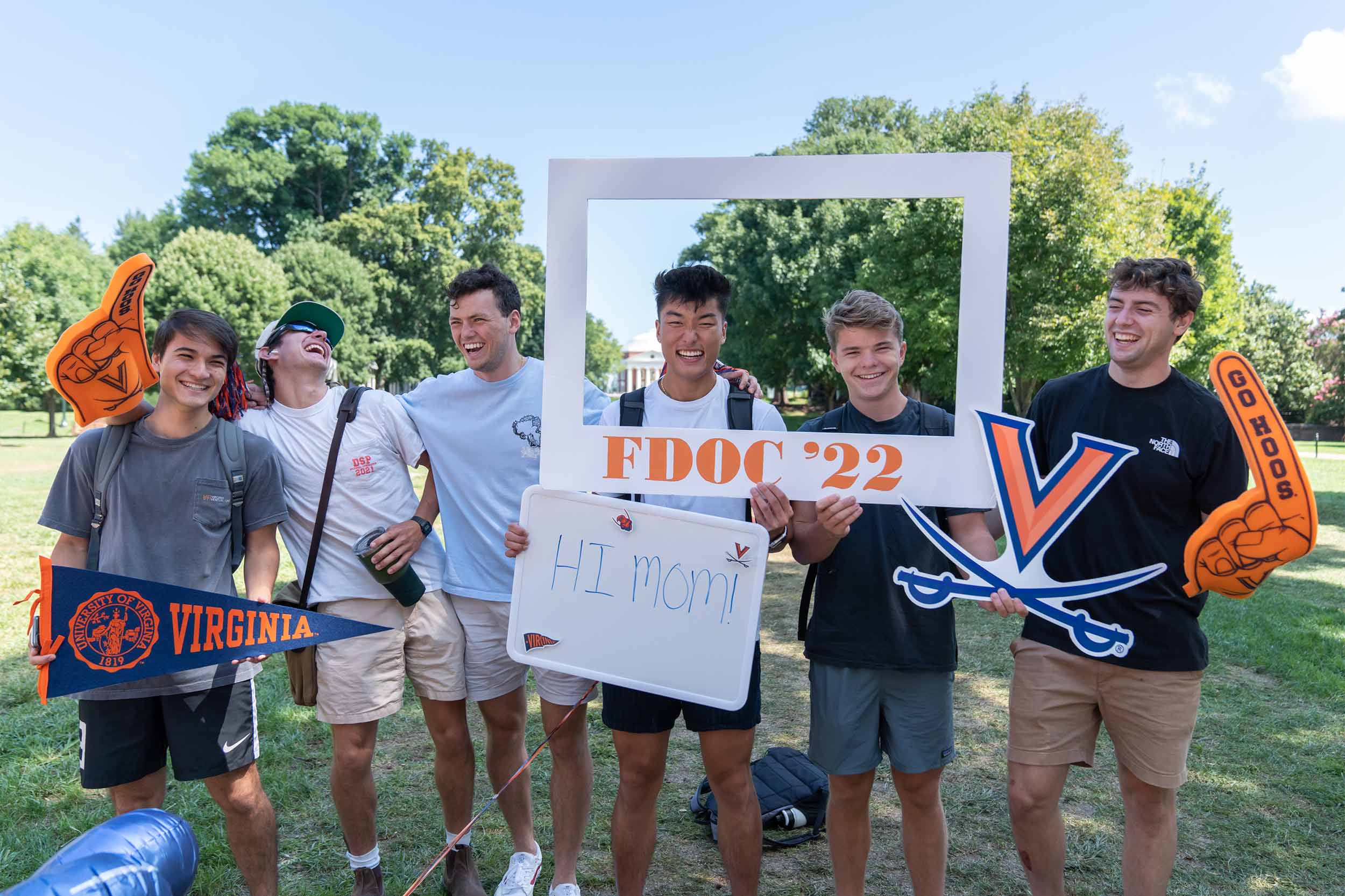 Six laughing students hold foam fingers, a Virginia banner, a whiteboard with Hi Mom written on it, and a frame reading FDOC '22