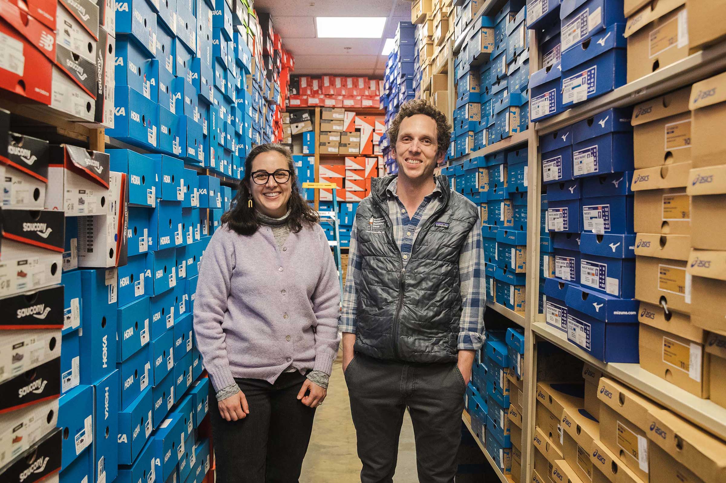 Audrey Sackson and Alec Lorenzoni standing in the running shop stock room, between two rows of shelves filled to the top with shoeboxes