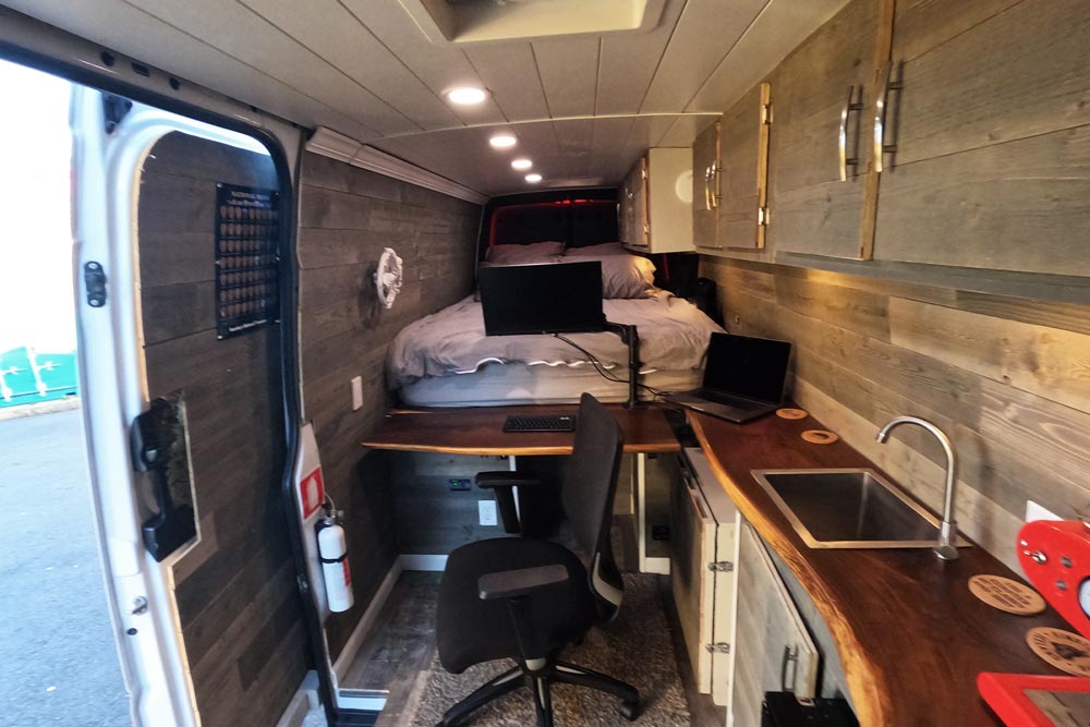 Interior view of the van showing a bed, a desk, chair, computer, sink, and the van door on the left.