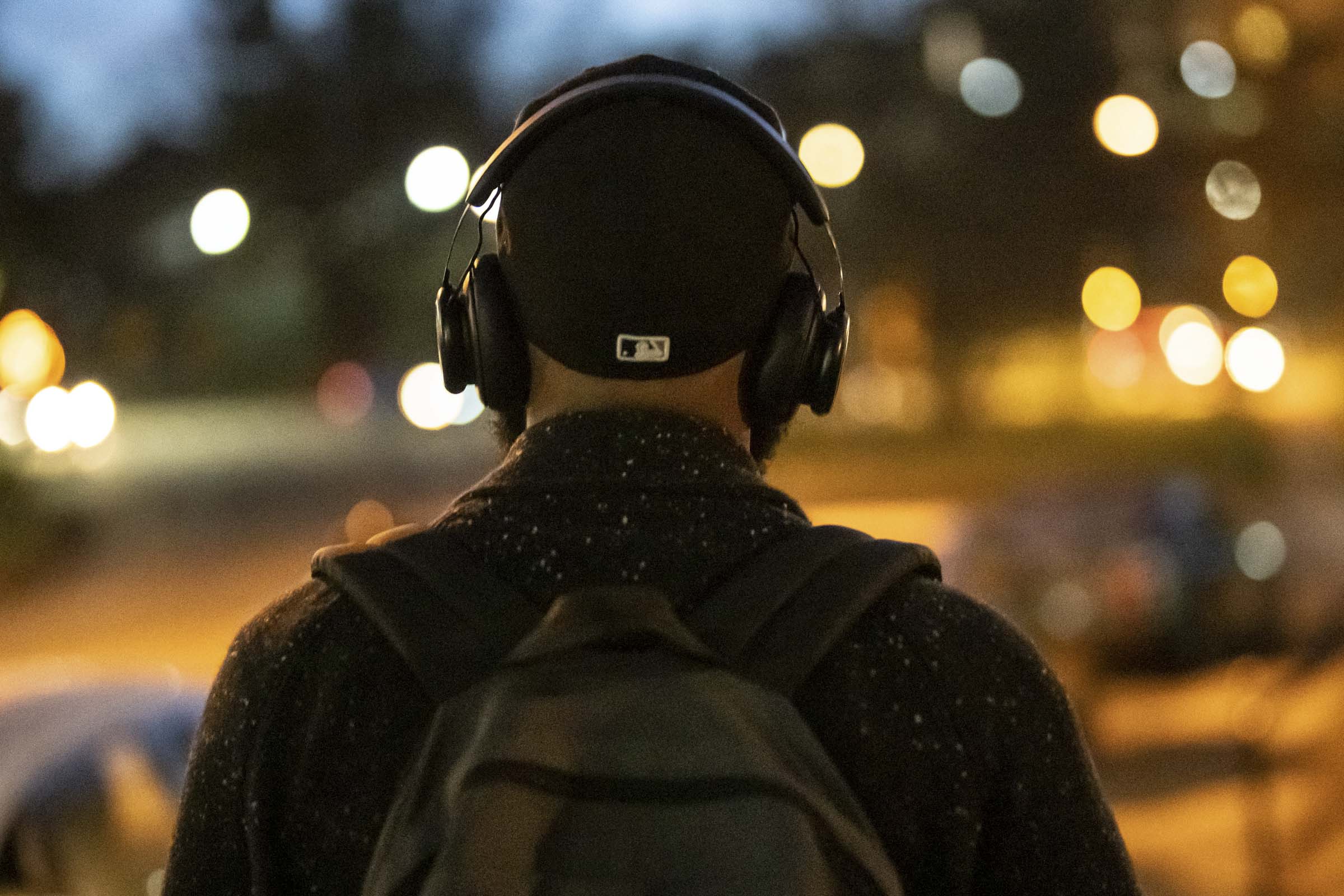 War lights twinkle in the street at dusk as Carson, wearing a backpack and headphones, walks away.