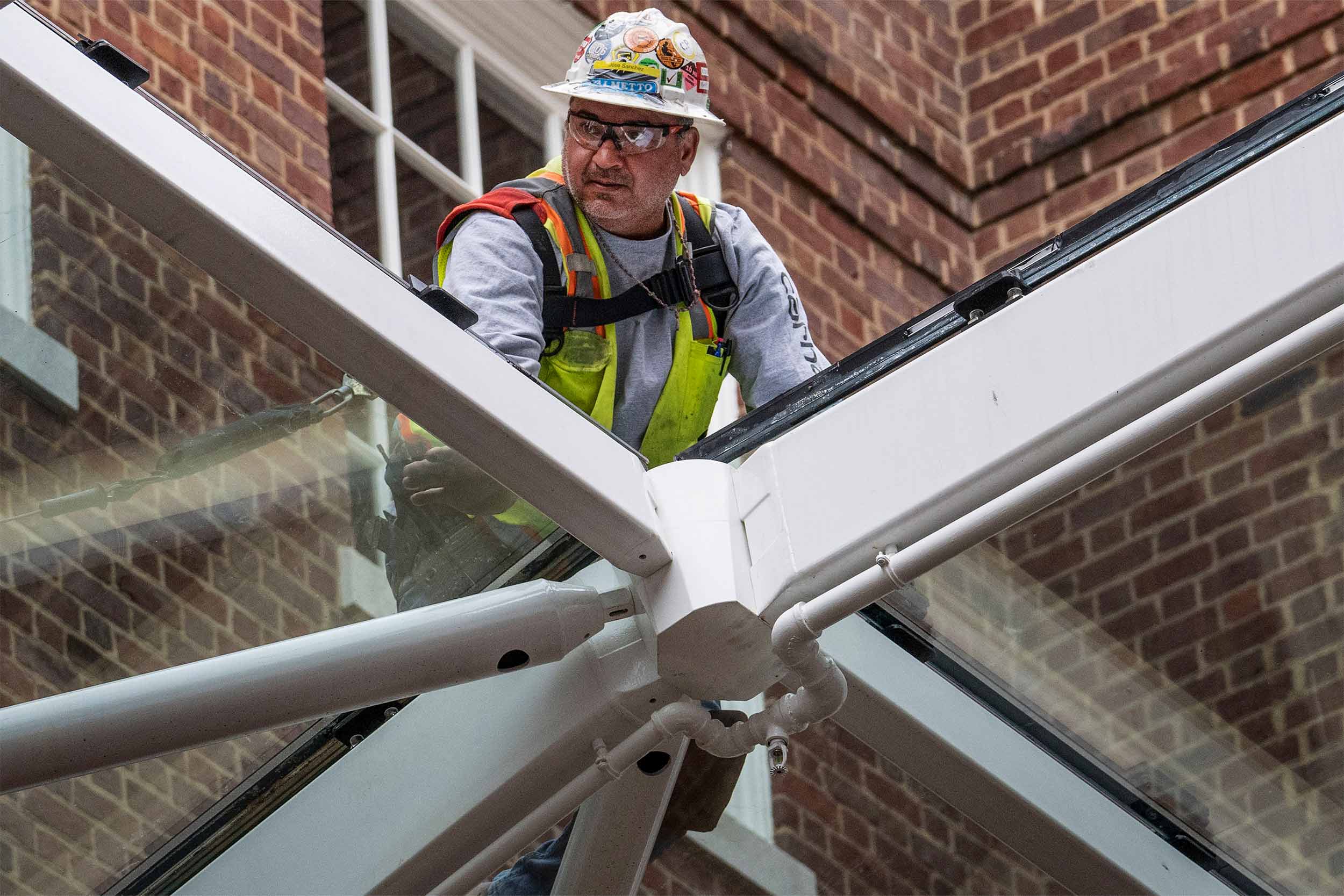 A worker on hands and knees on a pane of already installed glass guides another pane into place