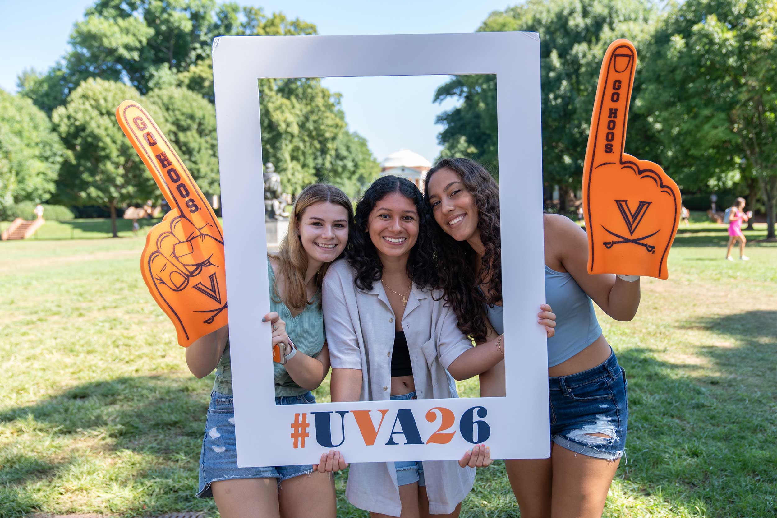 Three smiling students wear foam fingers and hold a #UVA26 frame