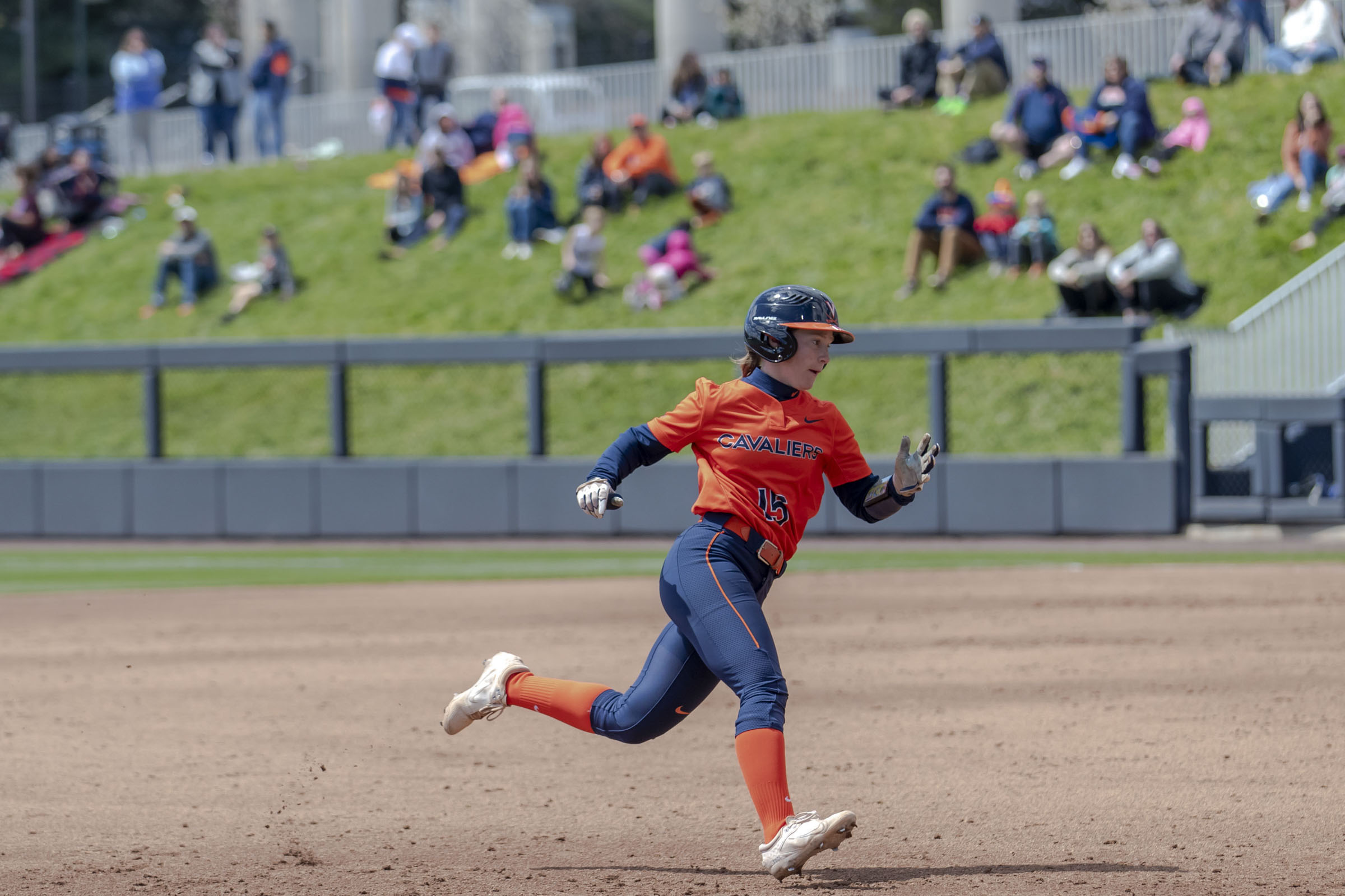 A UVA softball player runs to second base. People sitting on a grassy hill look on.