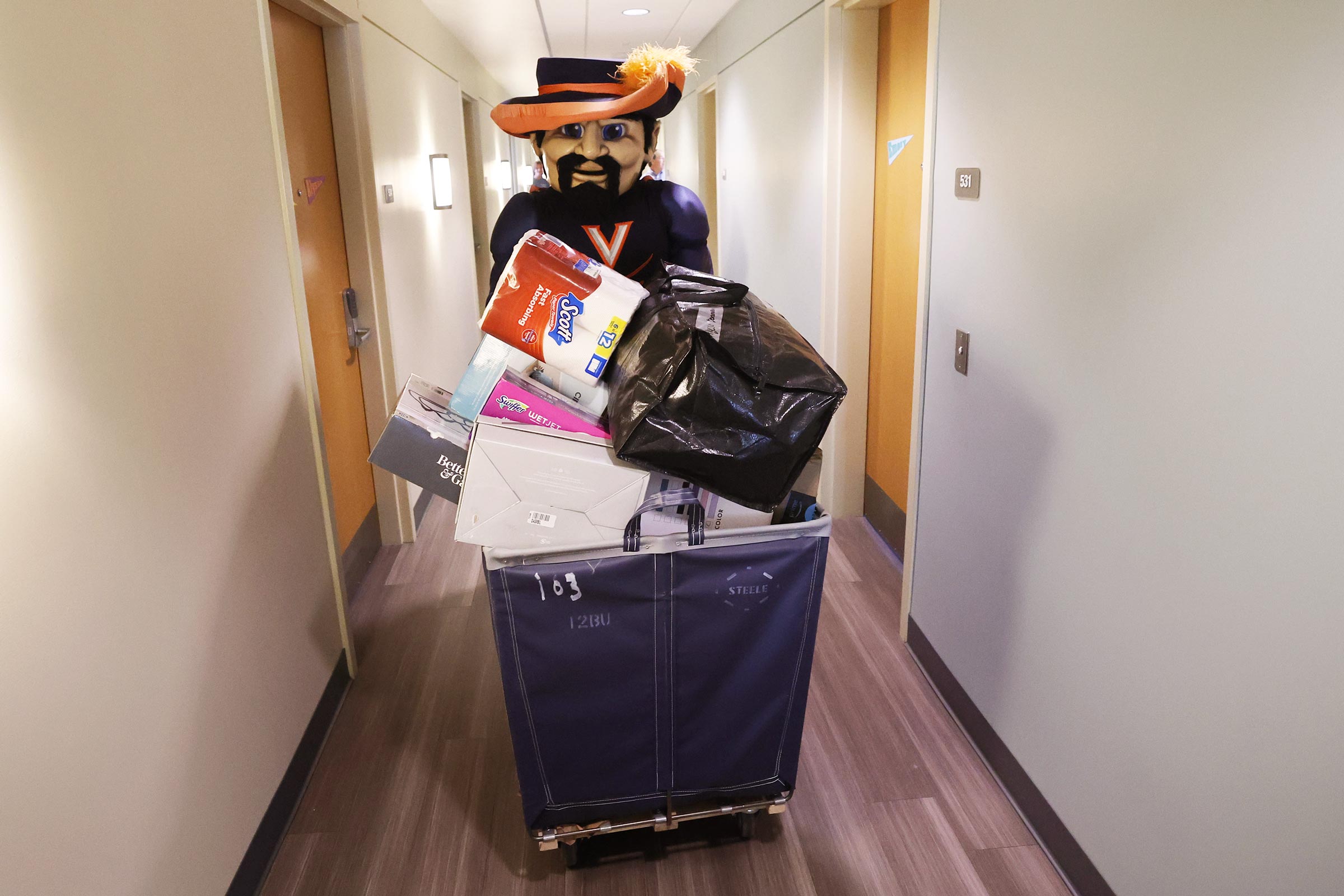 Cavman walks down a dorm hall approaching the camera with a cart of belongings