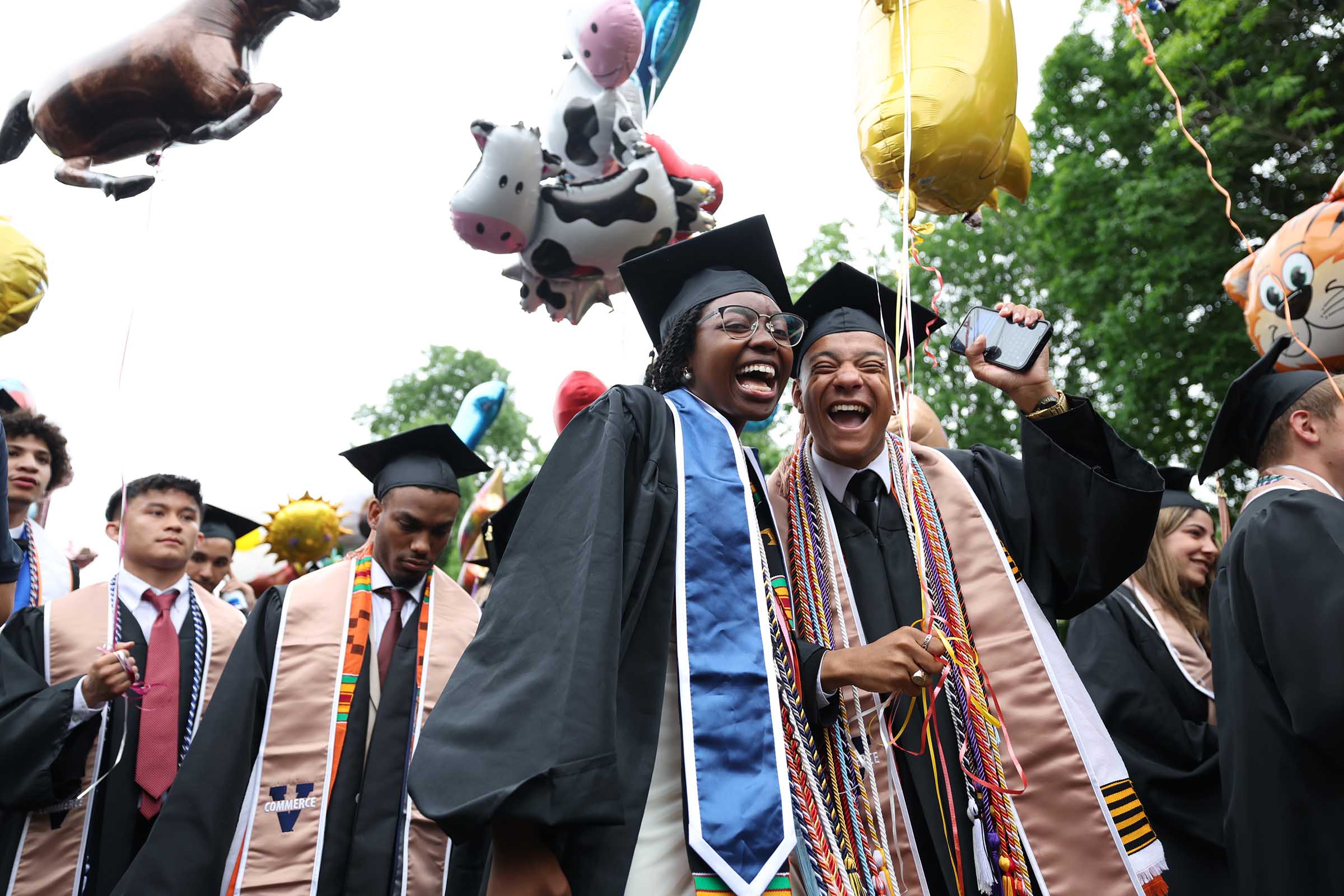 Two grads smile enthusiastically while holding their balloons along with several other grads in a crowd