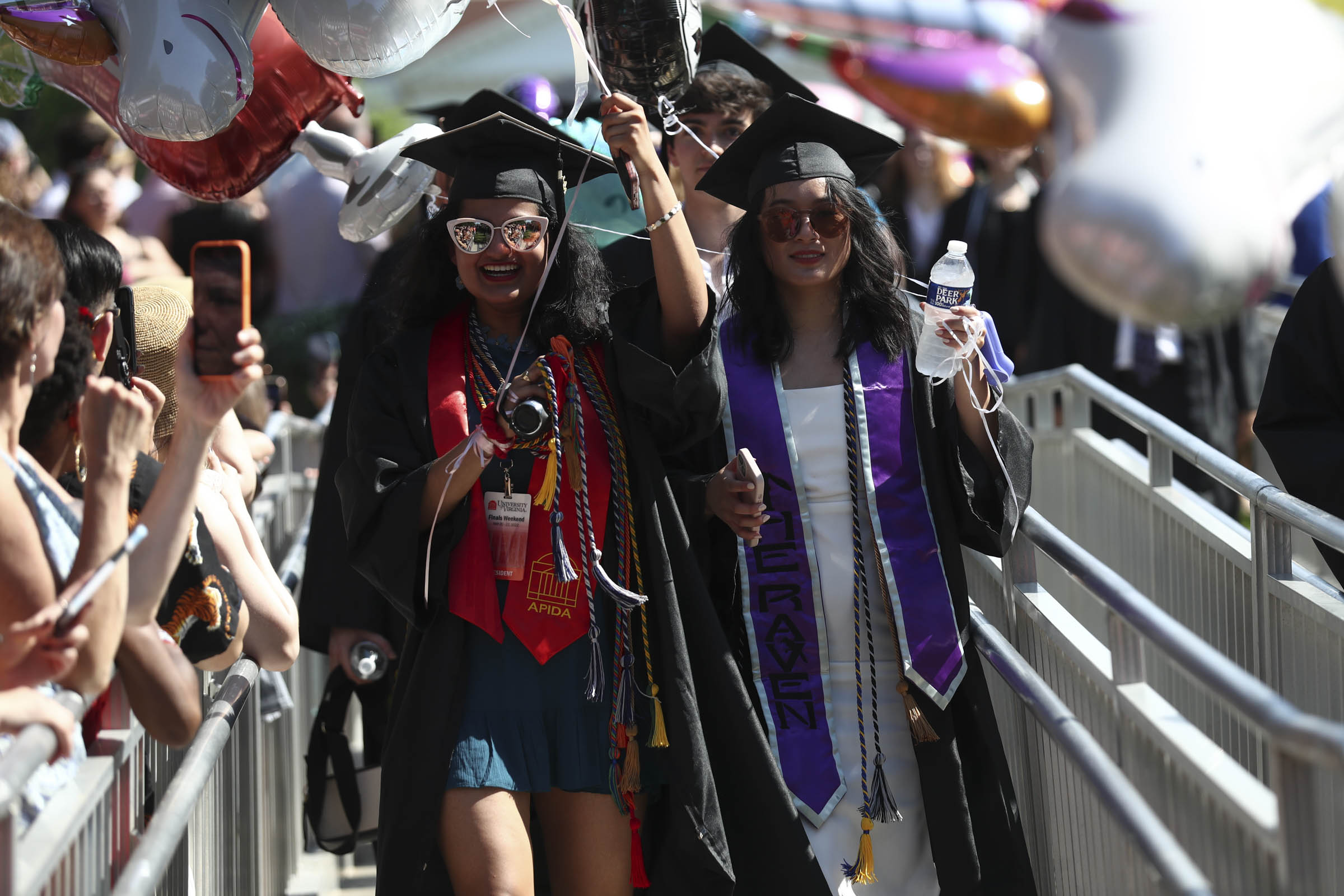 Two students carrying bottled water and balloons walk past a cheering crowd.