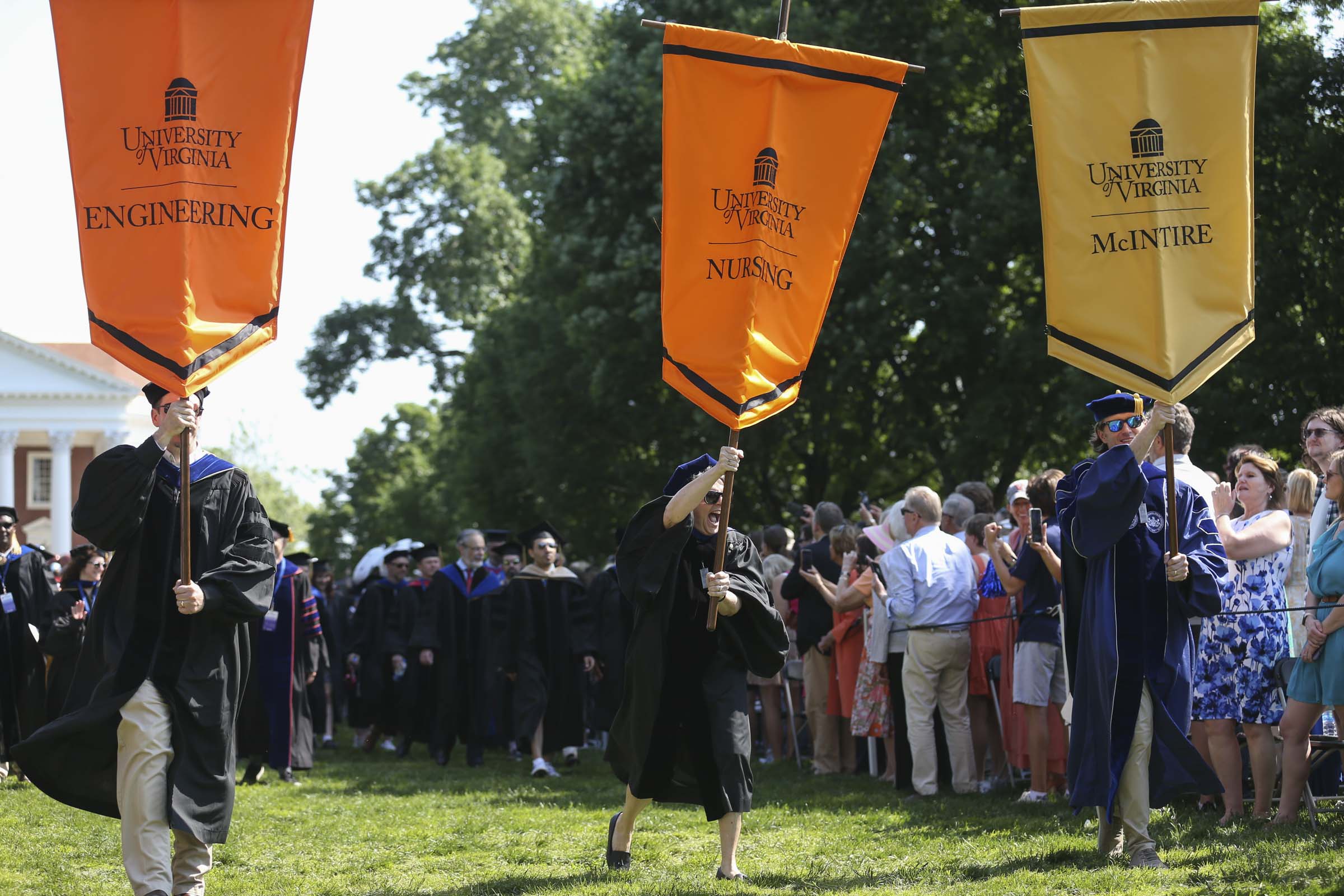 Standard-bearers hoisting banners for the schools of Engineering and Nursing, and the McIntire School, lead lines of their graduates across the Lawn.