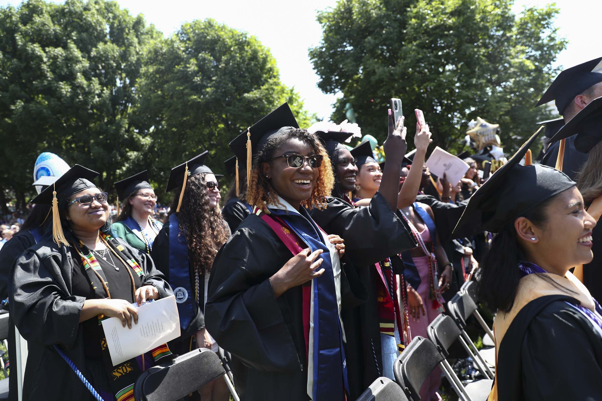 A student beams a smile and shows off her graduation sashes while raising her hand.