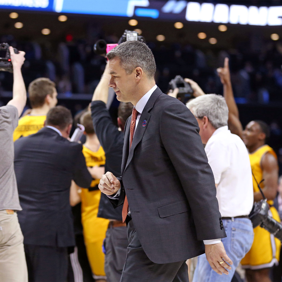 Tony Bennett walking after the court after UVA’s defeat with the UMBC team celebrating in the background.