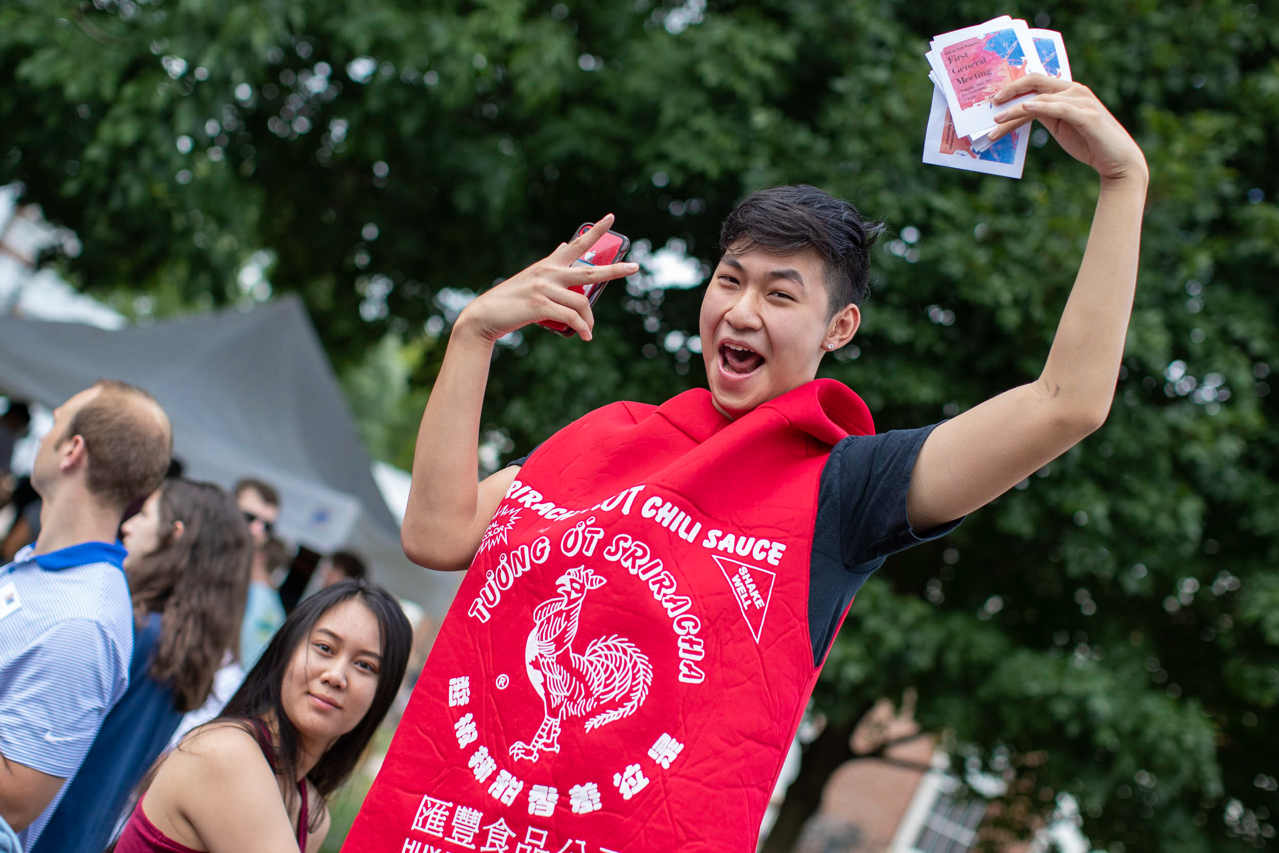 Student dressed in a Sriracha bottle costume holds fliers up and smiles at the camera