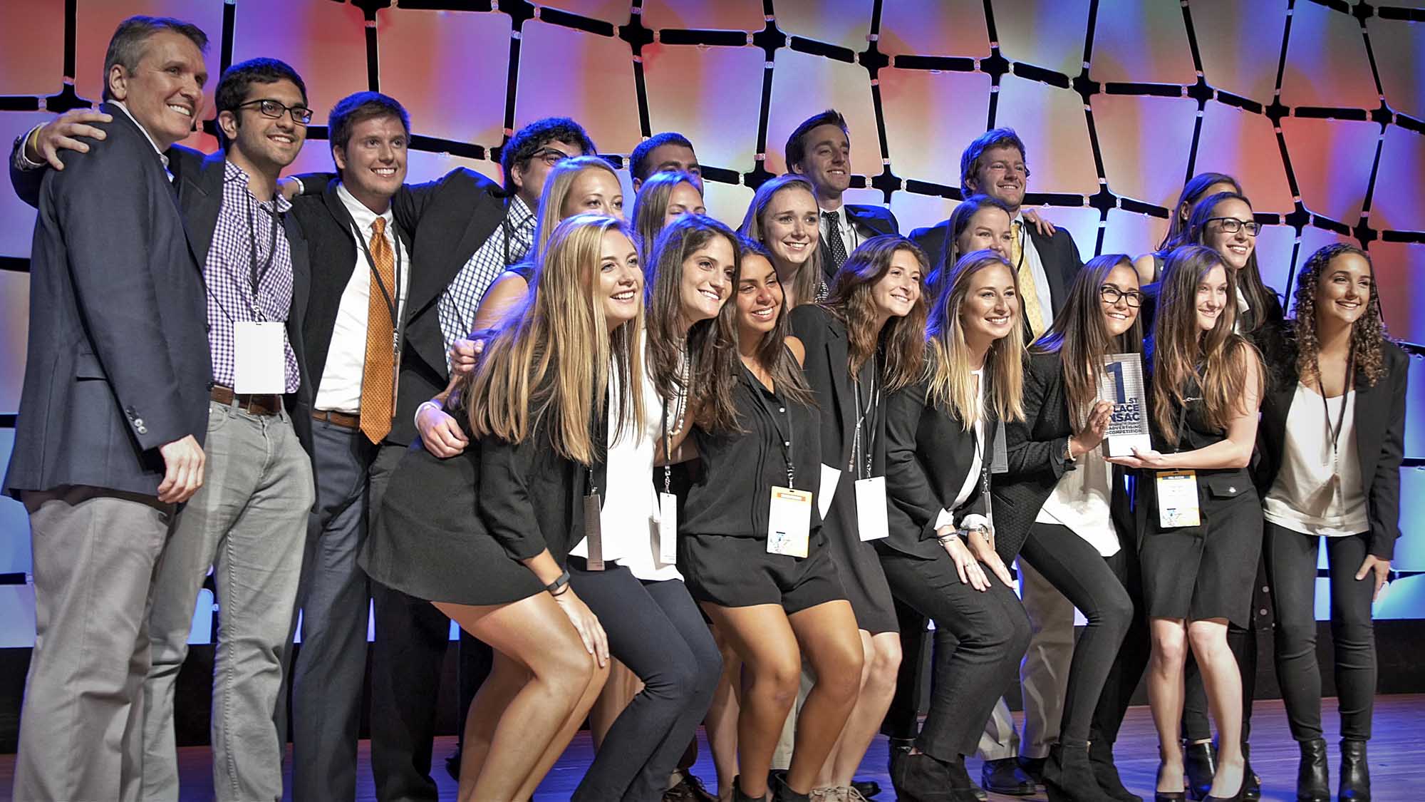 UVA Competitive Ad team pose together for a group photo on stage while they hold their first place trophy