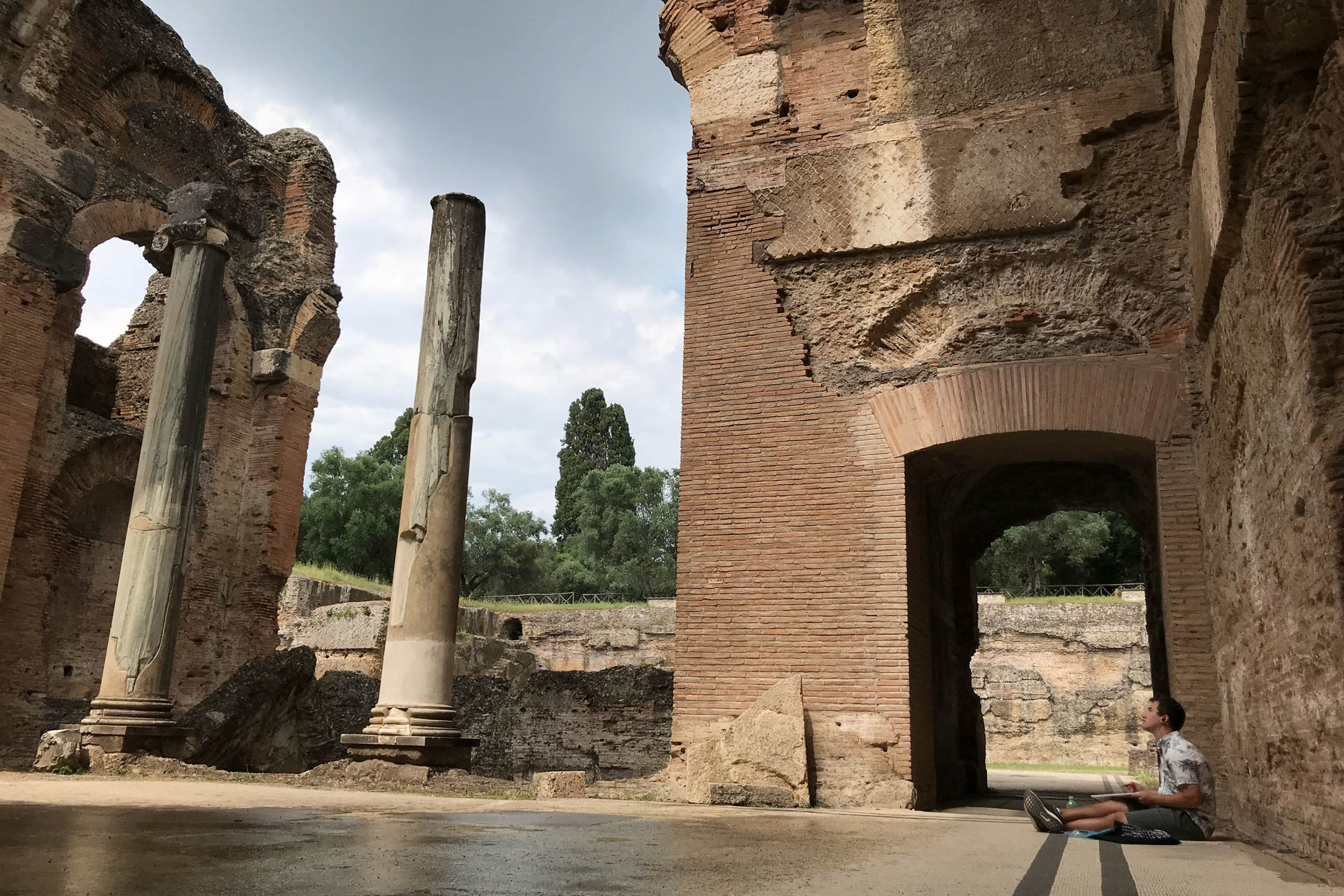 Michael Tucker sits on the ground sketching a roman ruin