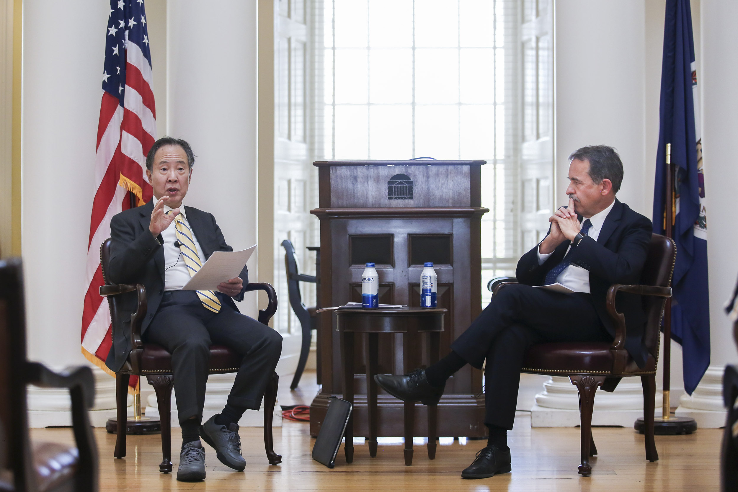 Koji Tomita, left, and Steve Mull, right, sit in two brown chairs while Mull looks at  Tomita speaking to a crowd