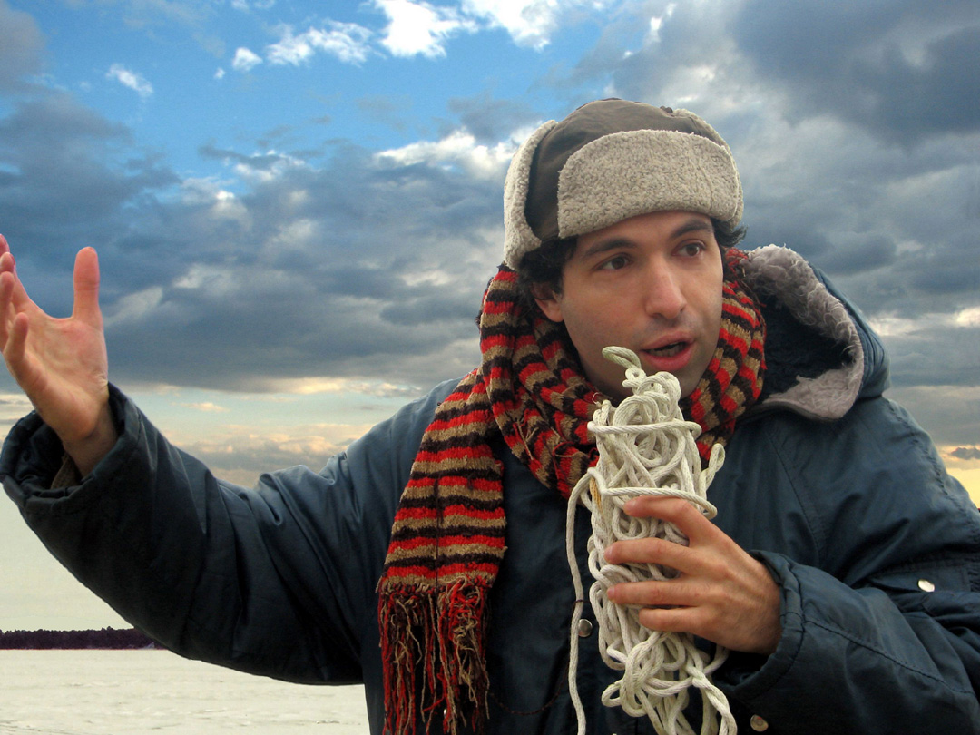 Man wearing a big coat, scarf, and hat holds a rope while talking