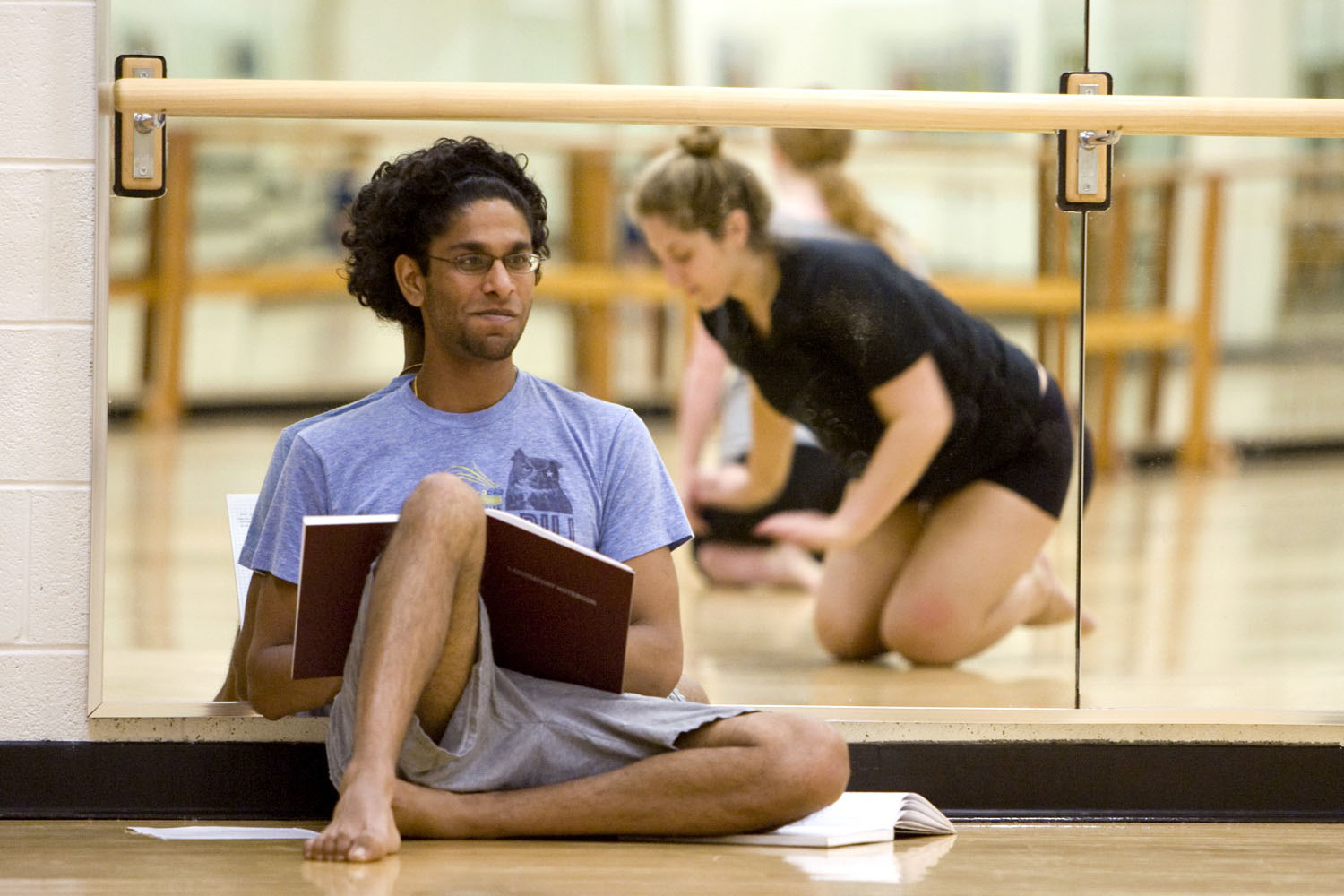 Shankar Srinivasan sits next to a glass wall with a notebook as he watches Rachel Swartz and Deidra Childressdance