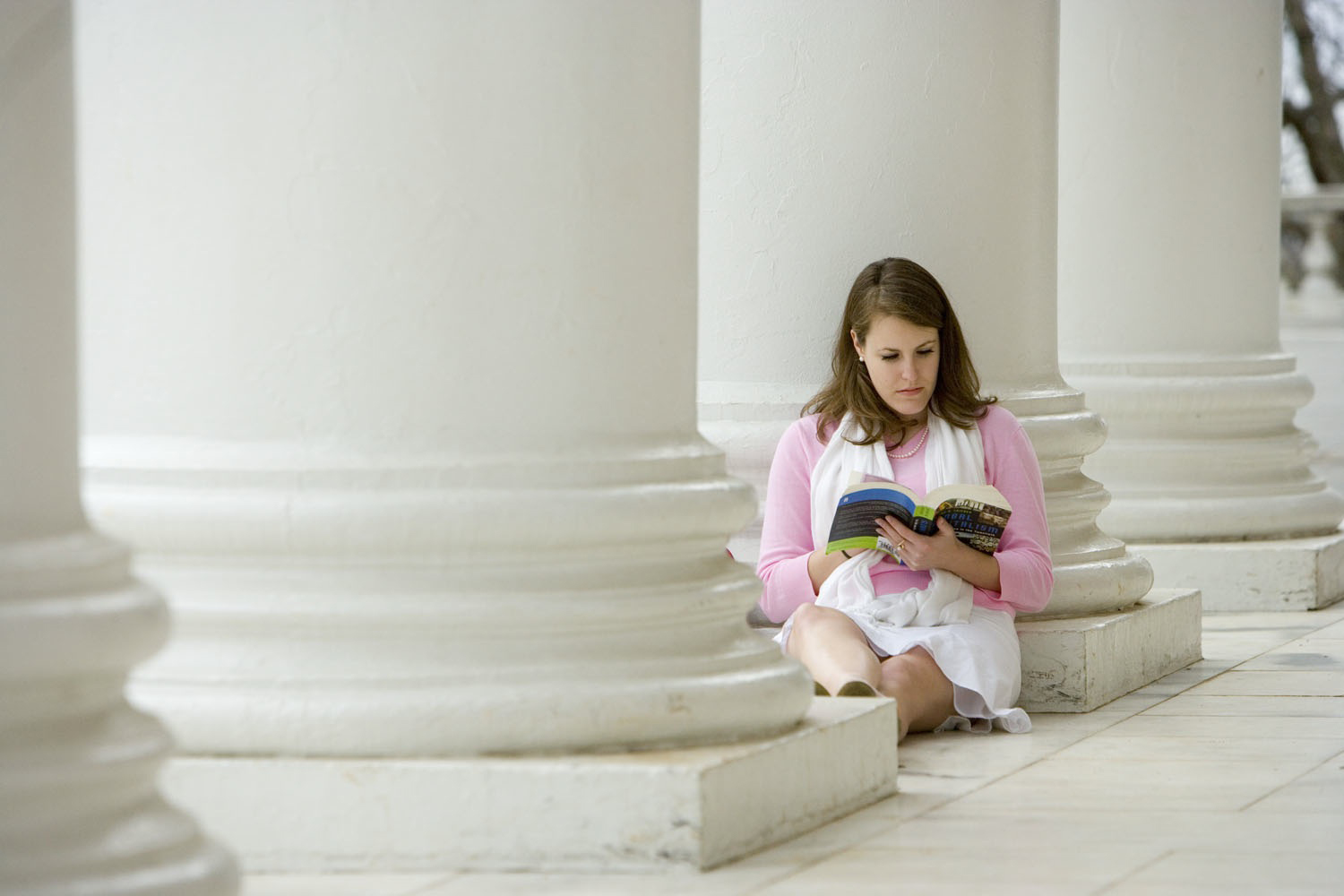 Woman sitting at the base of a white column reading a book