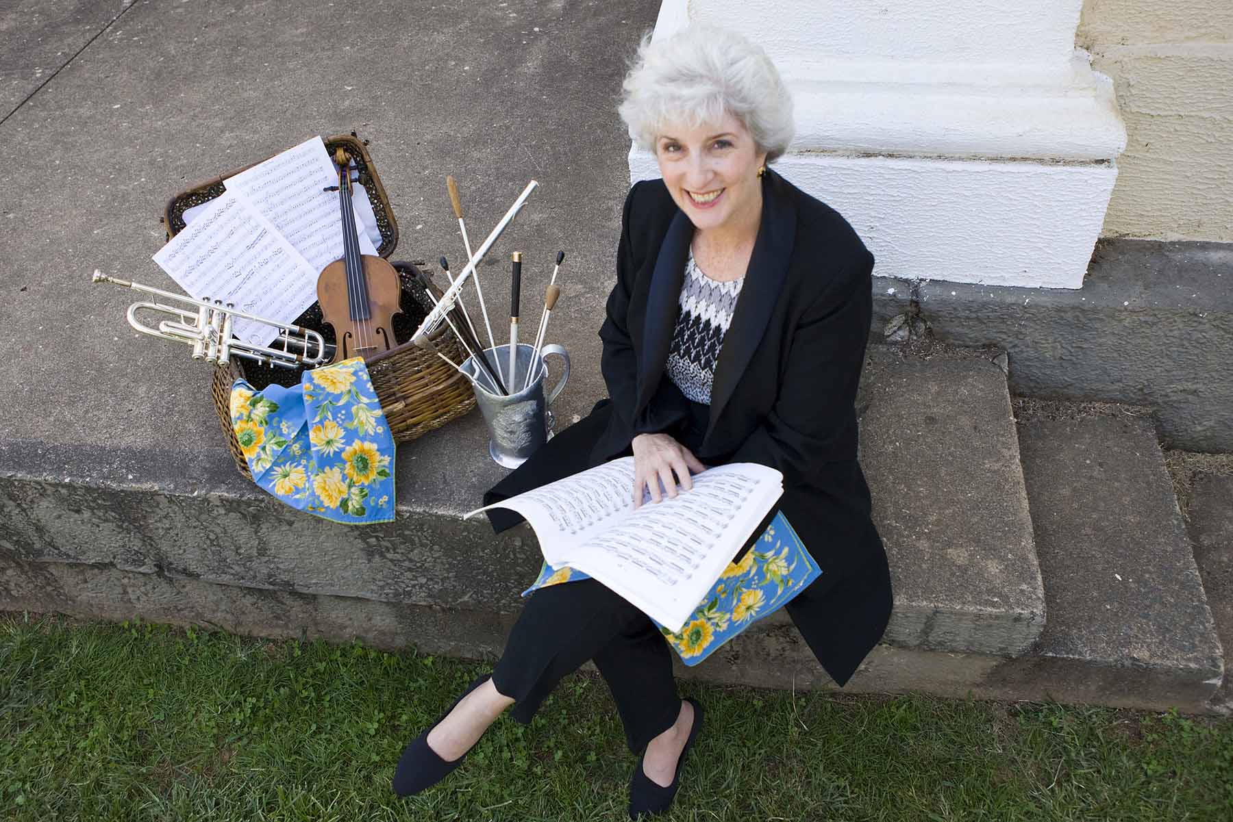 Kate Tamarkin sitting on concrete steps holding a music book with musical instruments in a wooden basket next to her