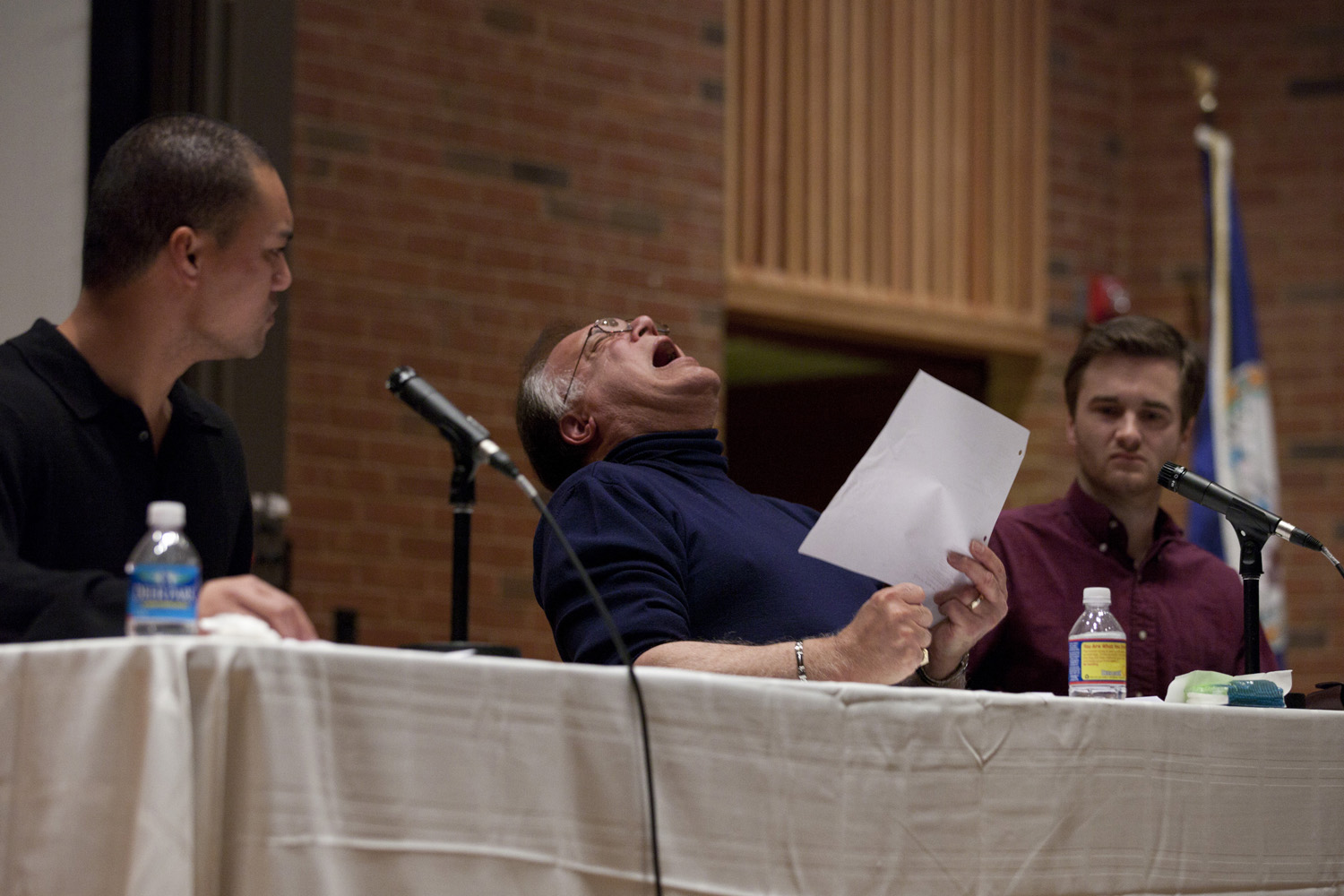 Men reading a script at a table for a crowd.  The man in the middle is in anguish as he puts his head back