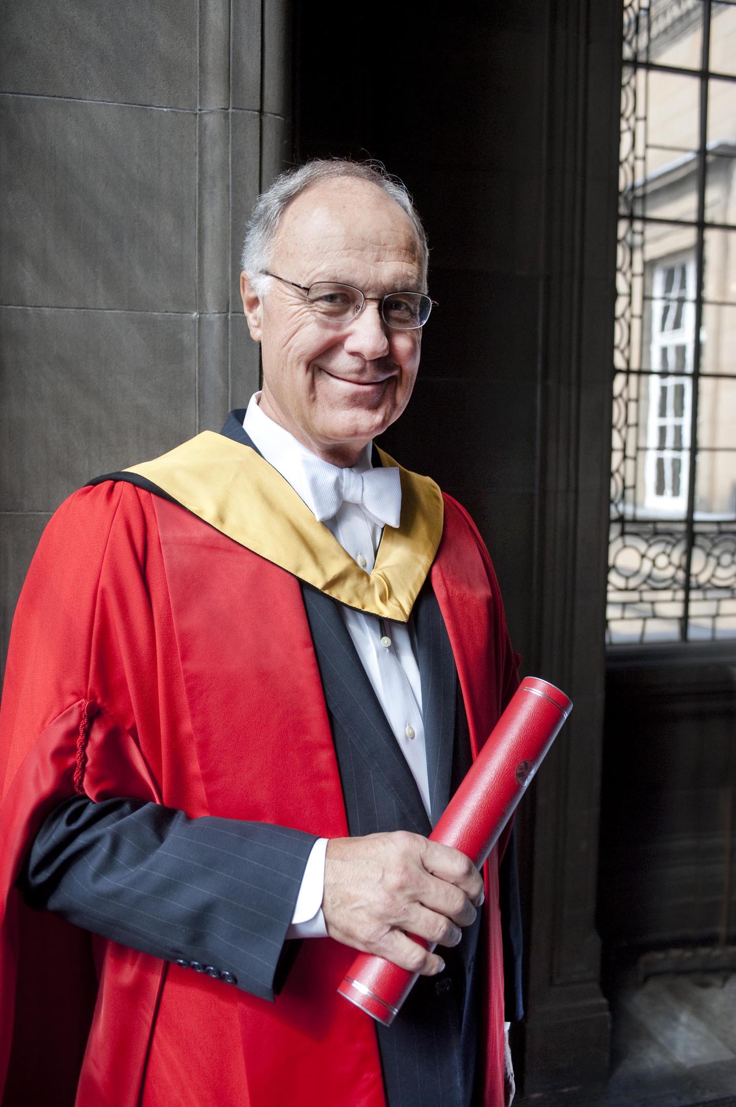 John T. Casteen III dressed in red graduation gown holding a diploma and smiling