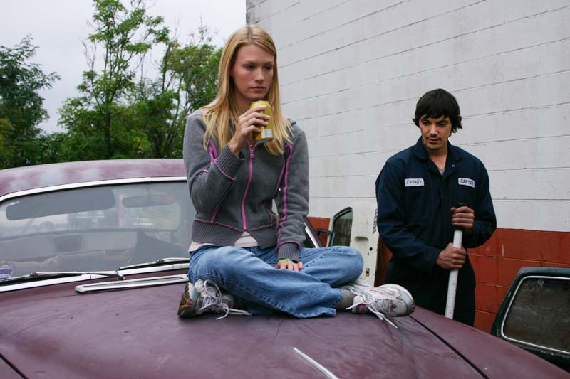 Woman sits on the hood of a car drinking a soda while a mechanic watches
