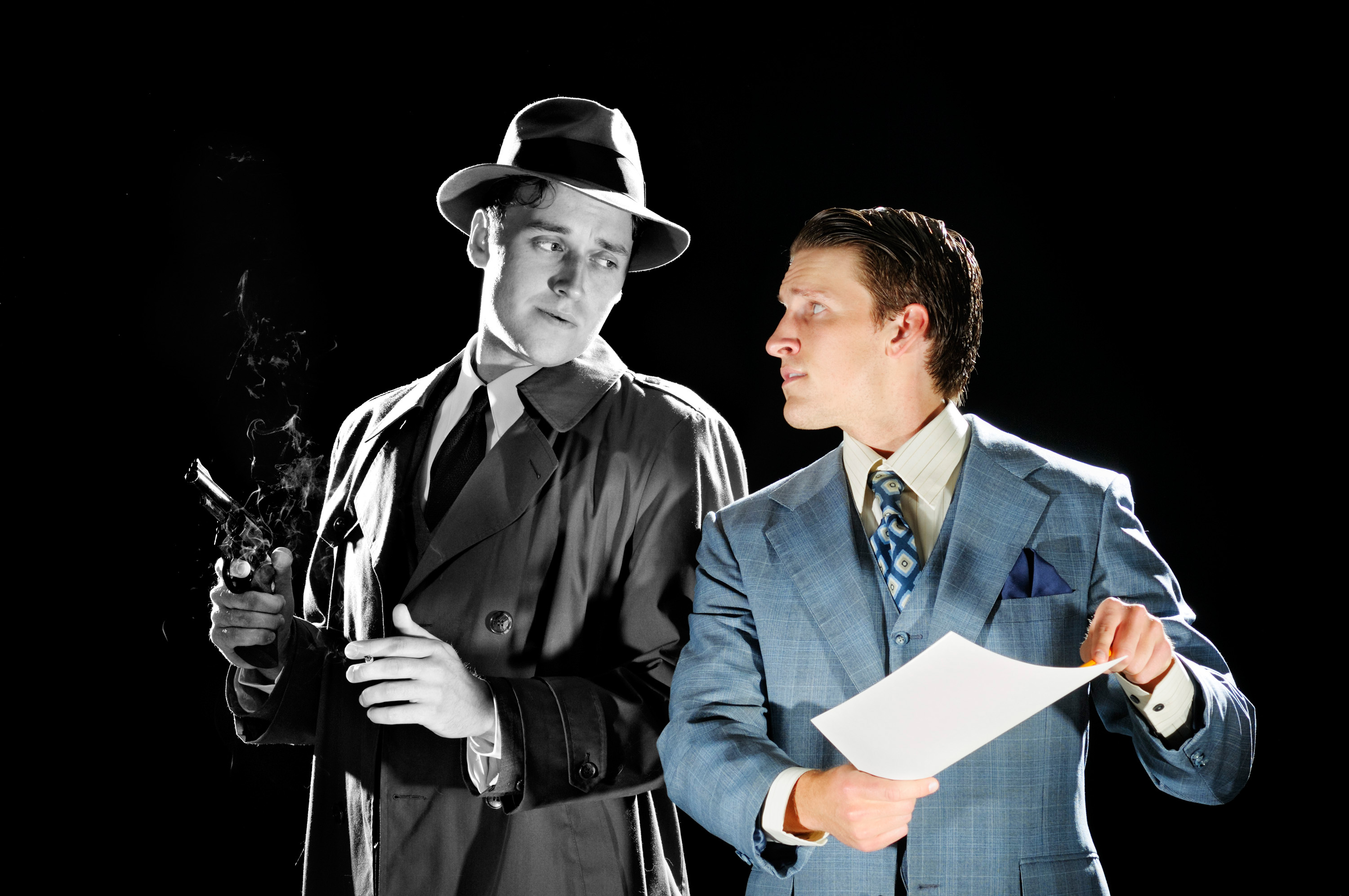 Two men standing on stage.  One is in a trench coat holding a gun the other is in a suit pointing at a piece of paper