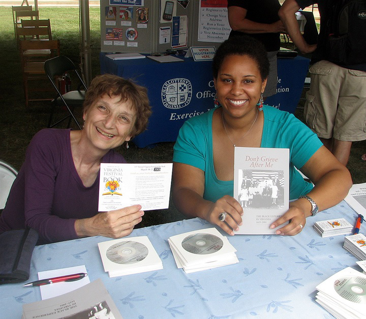 Nancy Damon left and Cameron Brickhouse, sit at a table holding a book and a flyer while smiling at the camera
