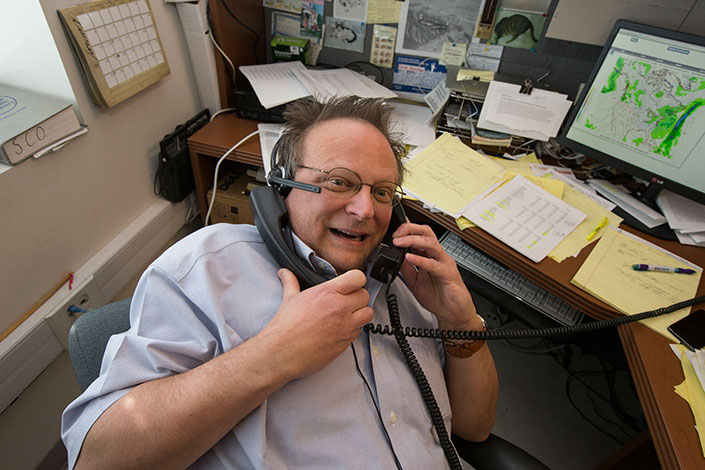 Jerry Stenger talking on two phones at a desk smiles at the camera