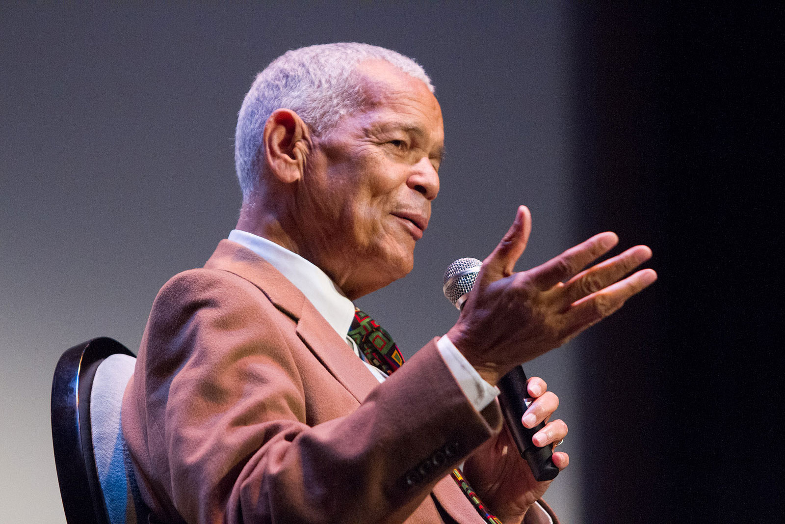 Julian Bond sits in a chair speaking into a microphone to a crowd