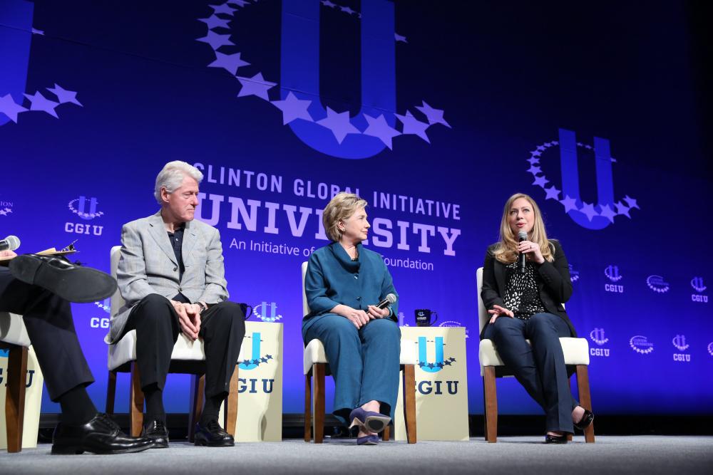 Bill Clinton, Hillary Clinton, and Chelsea Clinton all sit on stage while Chelsea talks to a crowd