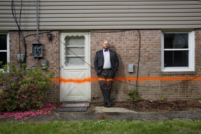 Man stands next to a brick house with an orange line spray painted across his legs and the wall