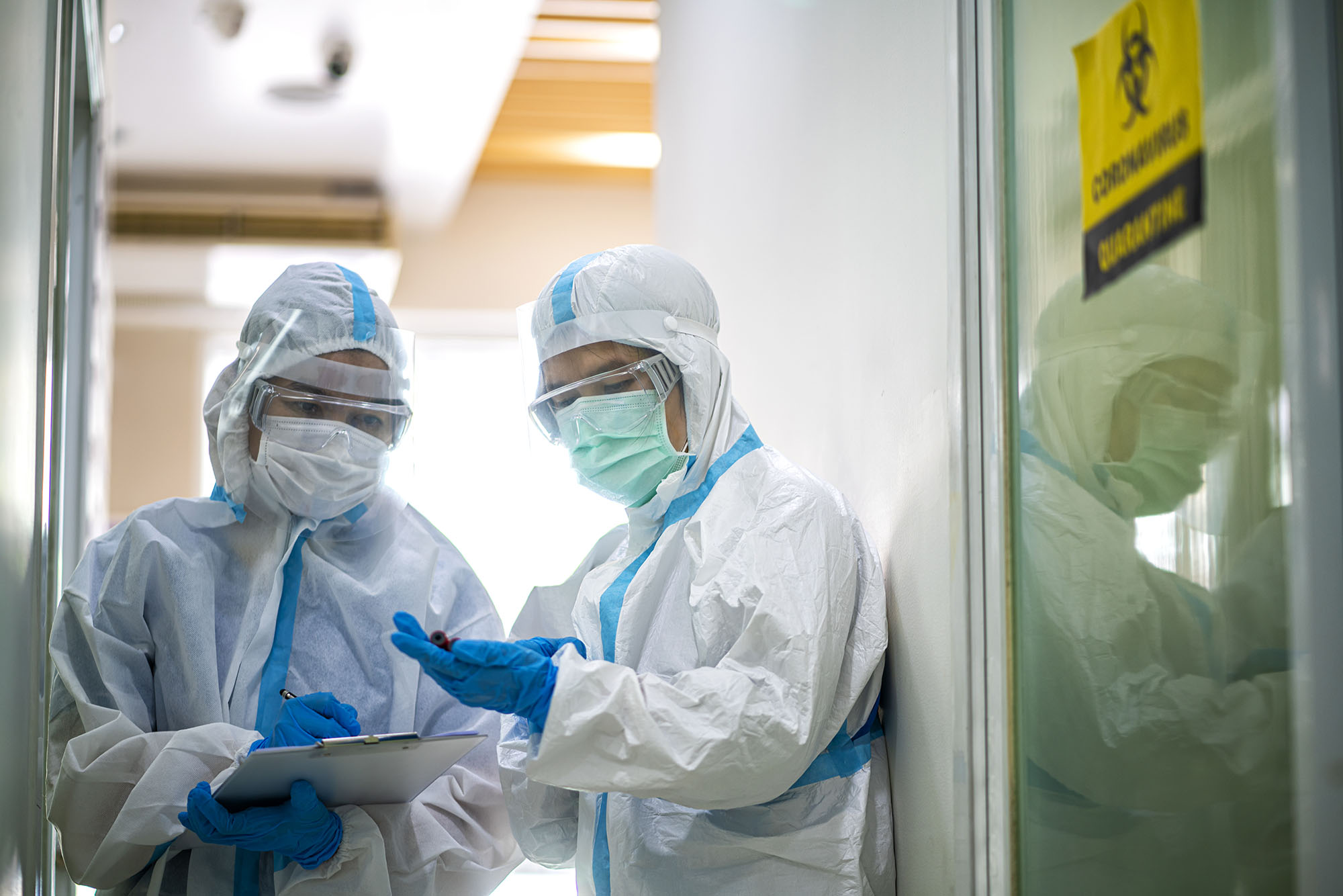 Two healthcare professionals in full PPE, one has a clipboard and pen, and the other is holding a tube that they are both looking at.