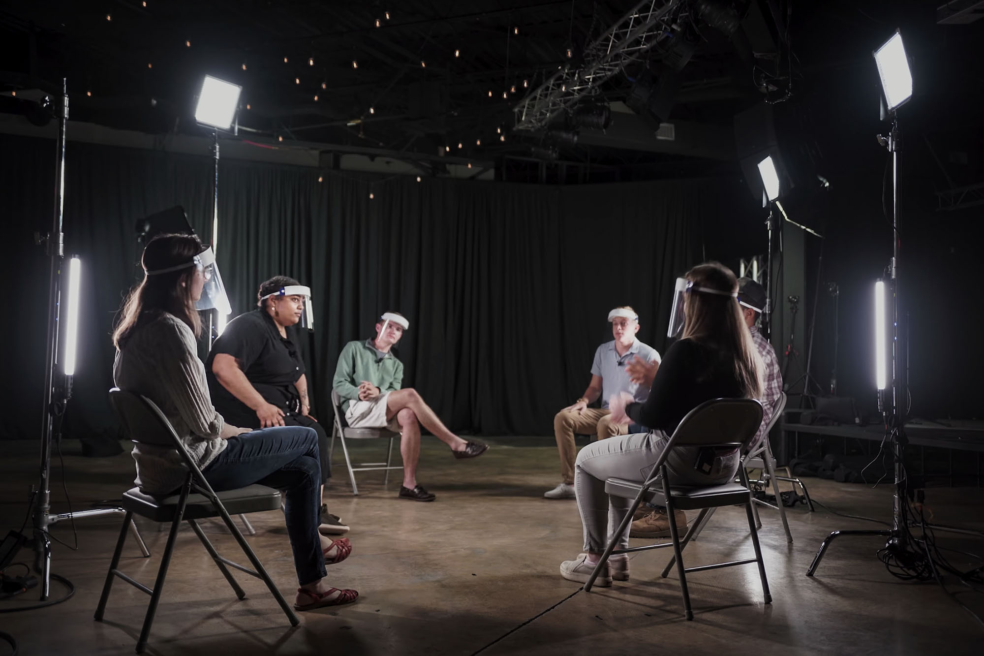 Group of people sitting in metal chairs talking to each other with bright lights for filming
