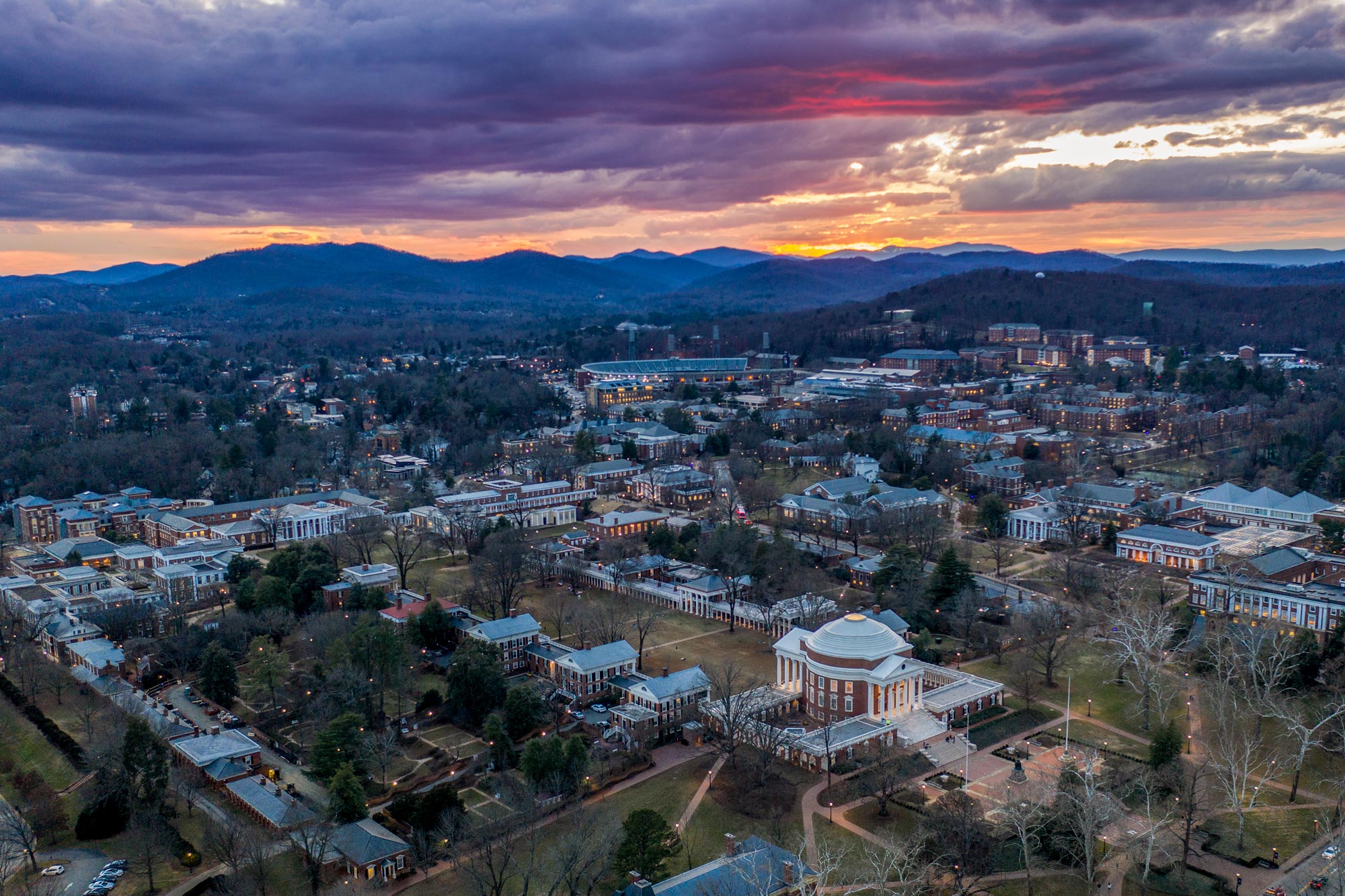 Arial view of the Rotunda at sunset with the sky, grey, purple, red, orange, yellow, and pink with the Blue ridge mountains in the distance