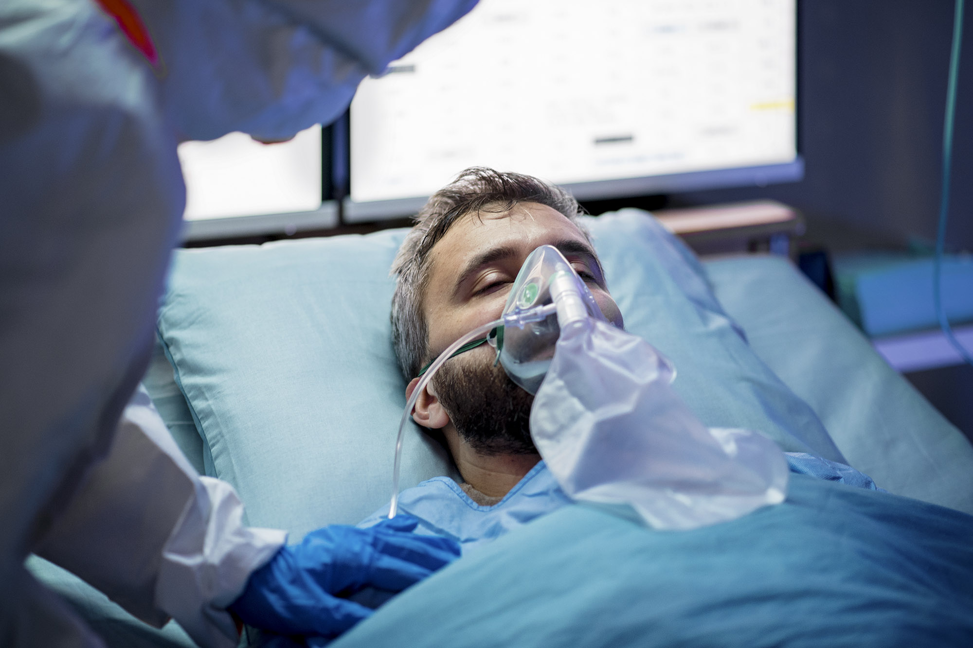 Patient laying in bed, with oxygen mask on their face with a medical professional standing over them