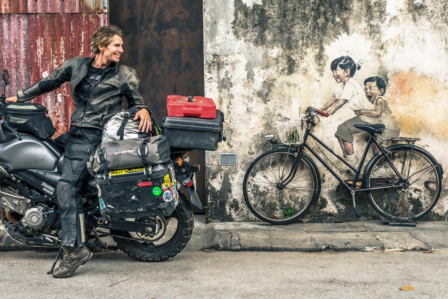 Eric Bernath stops his motorcycle on a Malaysian street to look at mural of two small children with a bicycle in front of it that looks they are riding it