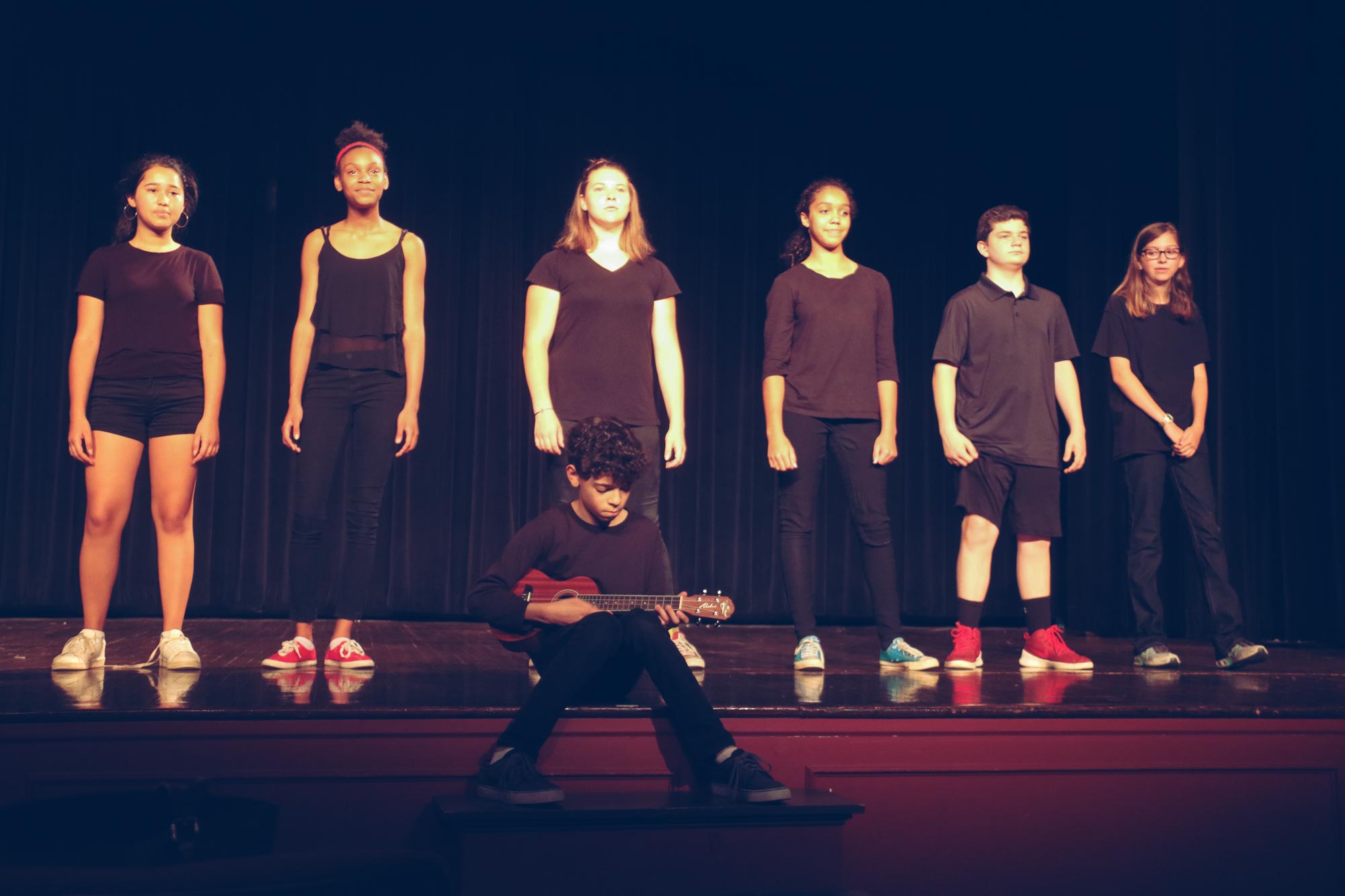Students stand behind one student who is sitting off the edge of the stage playing a guitar during a play