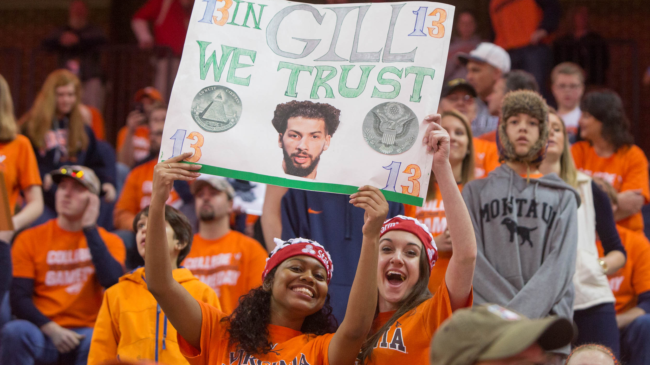 Two women at a basketball game holds a sign that says in Gill we trust