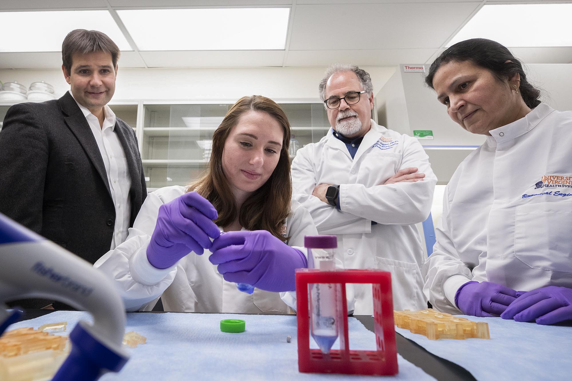 People standing behind a woman who is sitting at a table watching her work with a test tube