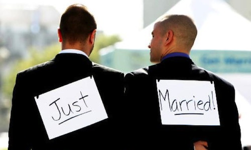 Two men stand holding an arm around each other with  a sign that reads Just married