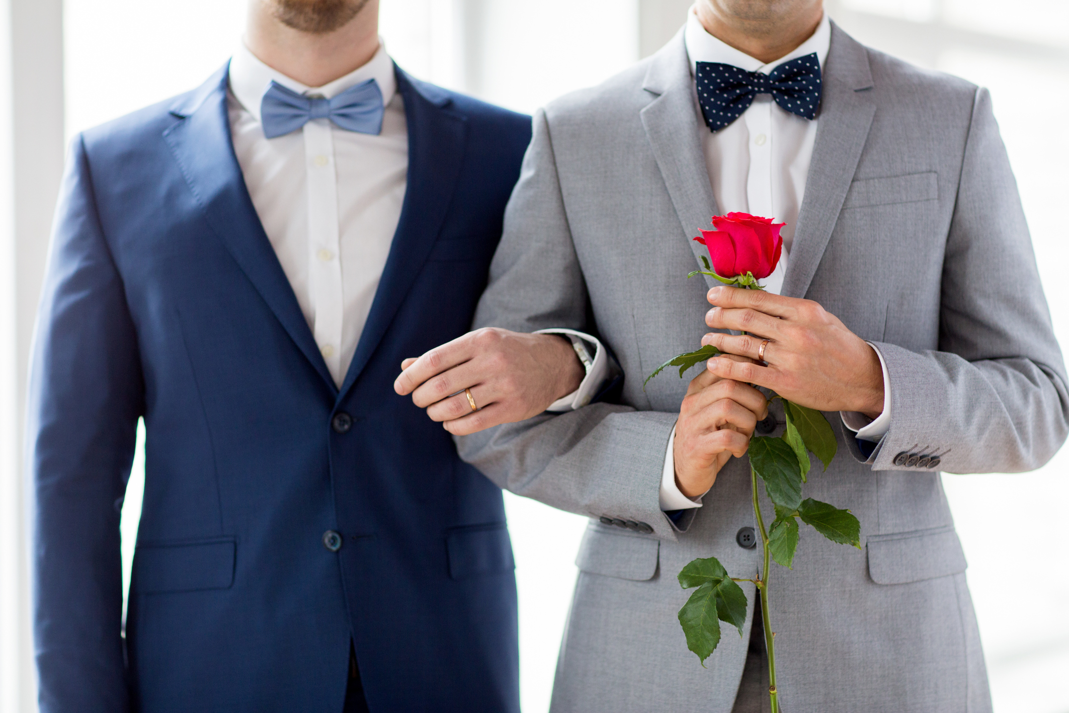 Two men standing next to each other in suits.  Both wearing wedding bands and their arms interlocked.  One man is holding a red rose