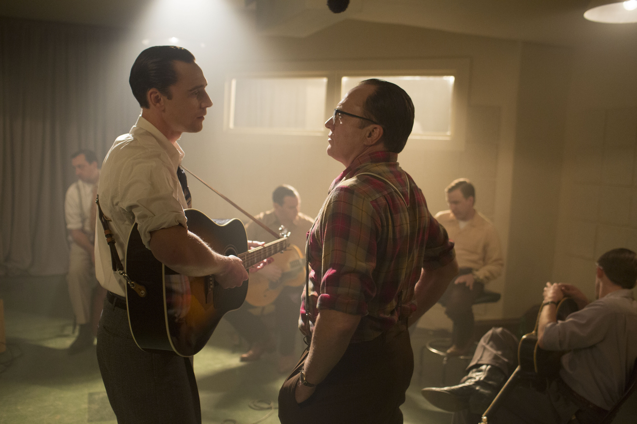 Actor playing a guitar looking at a man standing directly in front of him.  Rest of the actors in the band are sitting listening
