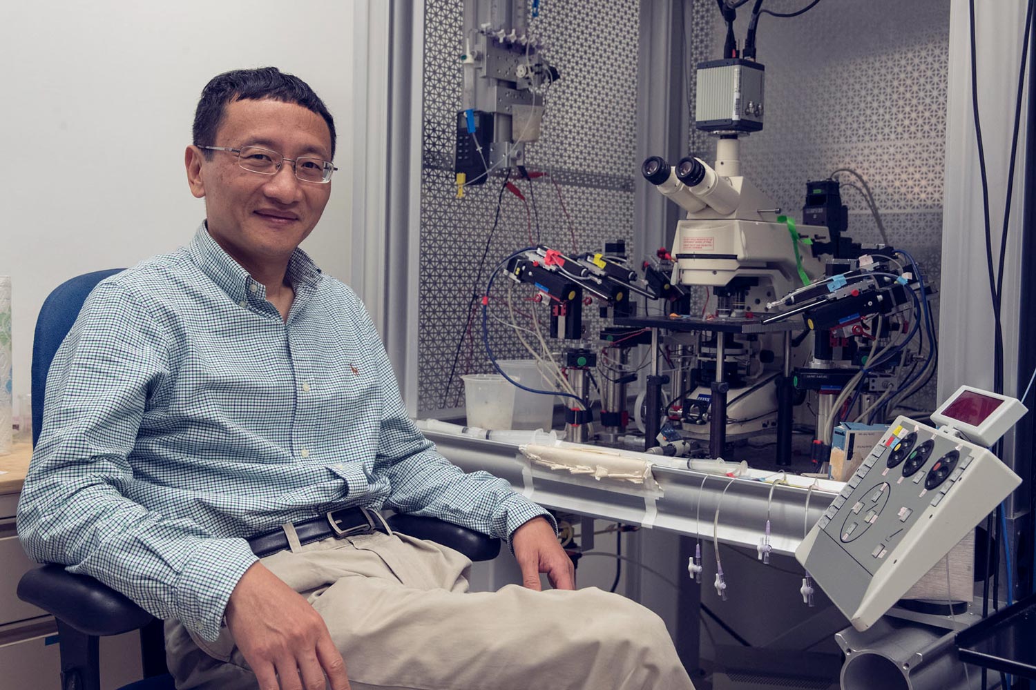 J. Julius Zhu sits in a chair next to a microscope looking at the camera