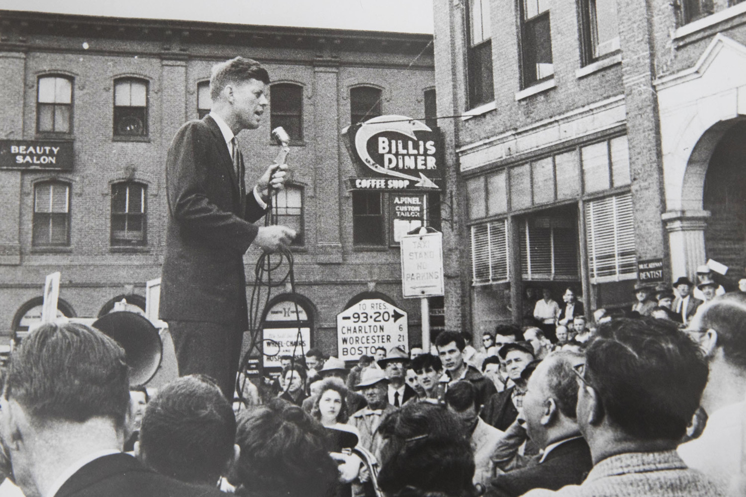 Black and White image of John F. Kennedy standing above the crowd in a street talking into a microphone