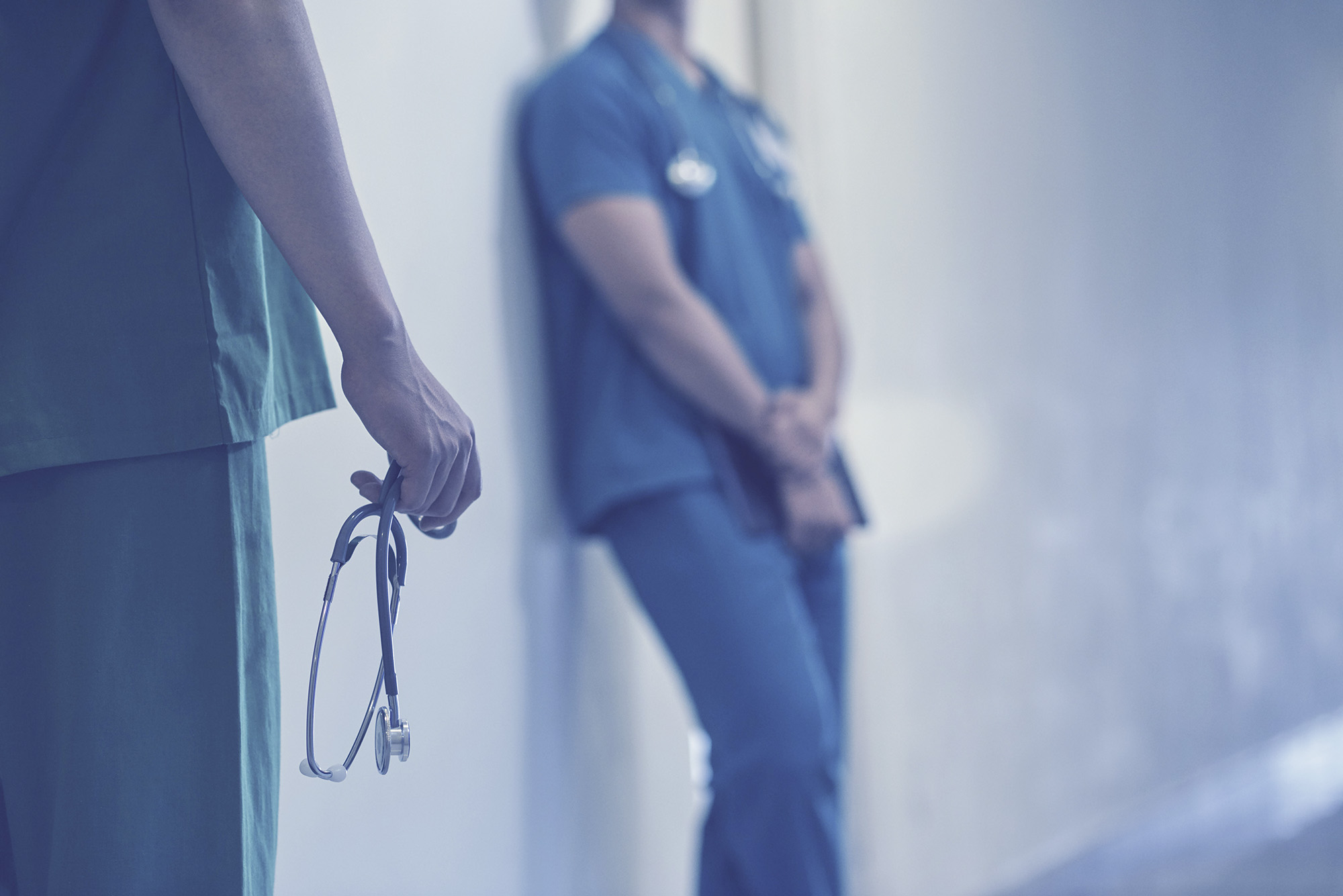 Medical Professional leaning next to the wall and one standing holding stethoscope 