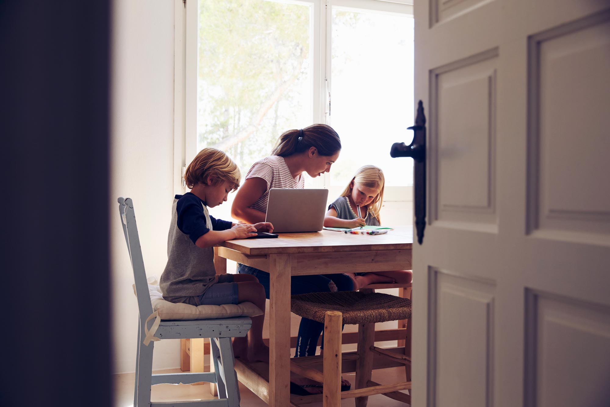 Woman sitting at a wooden table helping two children sitting on both sides of her with their homework