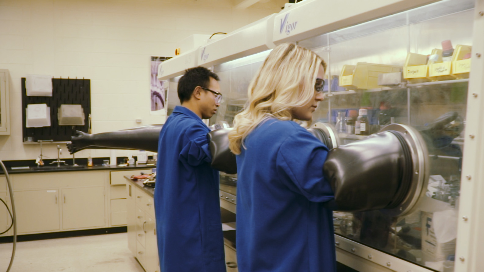 Two researchers putting their arms into rubber gloves to work in a contained environment