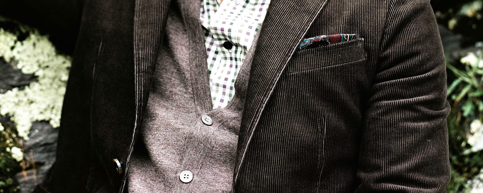Up close of a man wearing a corduroy jacket, grey button up vest and a plaid dress shirt