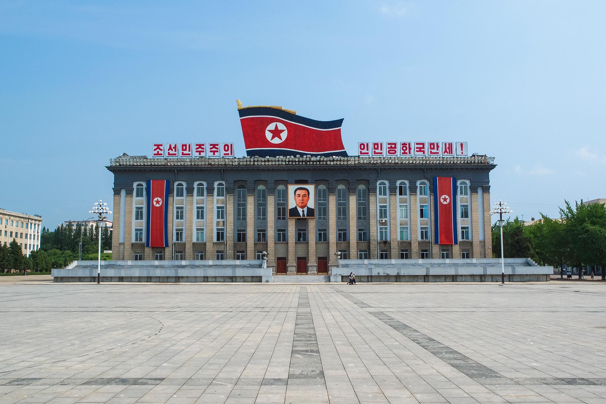 Kim Il Sung Square in Pyongyang, North Korea,  is a four story building with a huge flag in the middle of the roof and a picture of him above the entrance